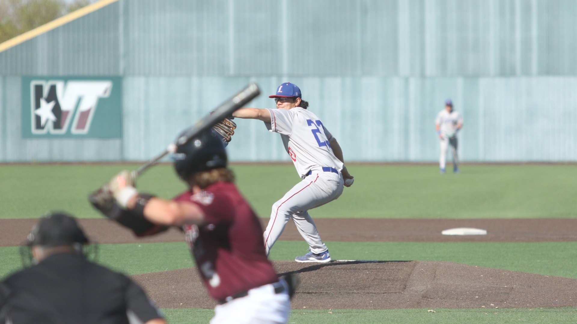 This is a photograph of Lubbock Christian pitcher Aaron Rubio (grey jersey, #23) throwing a pitch to a West Texas A&M batter at Wilder Park.