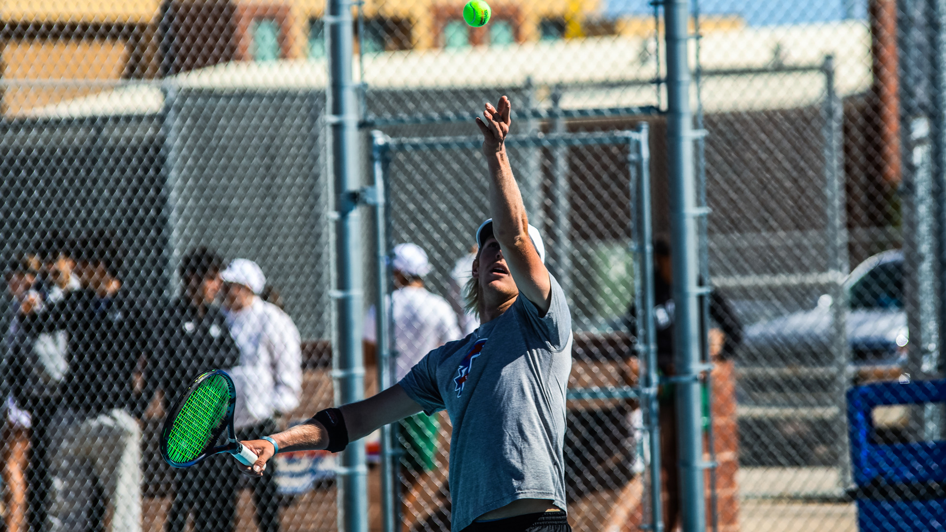 An LCU men's tennis player in a gray LCU shirt tosses the ball upward to serve, with teammates and spectators visible in the background behind a chain-link fence.