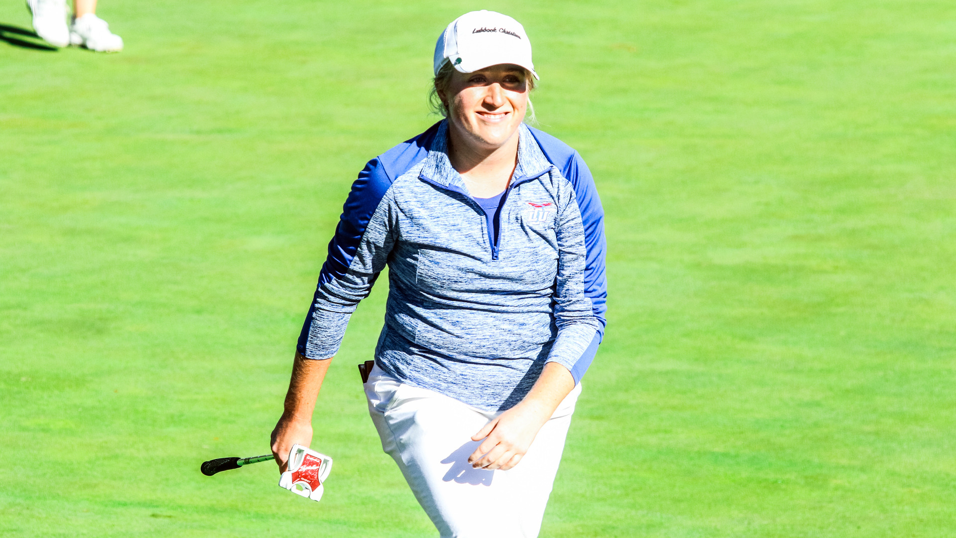 A smiling LCU golfer in a blue and gray LCU quarter-zip pullover and white Lubbock Christian cap walks a sunlit fairway carrying a putter.