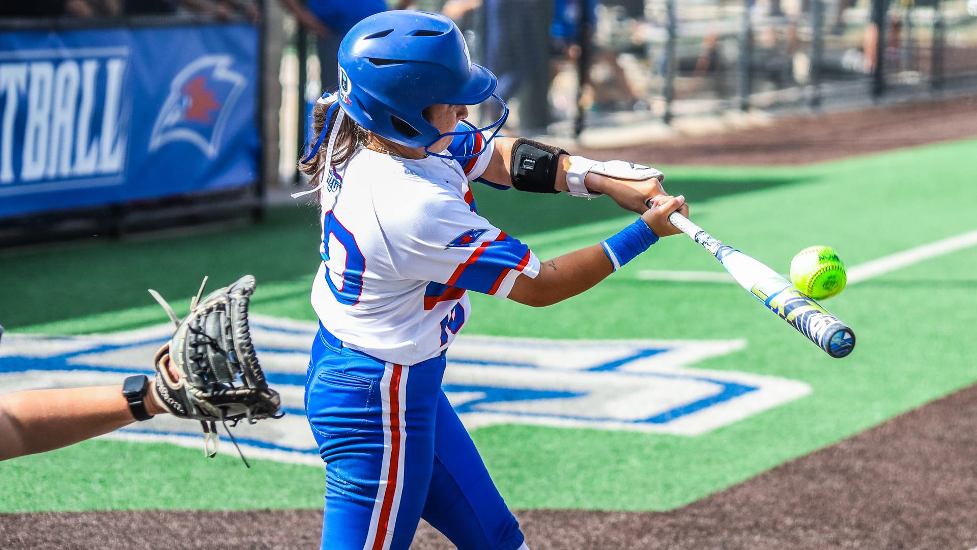 An LCU softball batter in a white and blue Chaps uniform and blue helmet makes contact with a yellow softball, with the LCU Chaparrals logo visible on the green turf and a blue LCU Softball banner in the background.