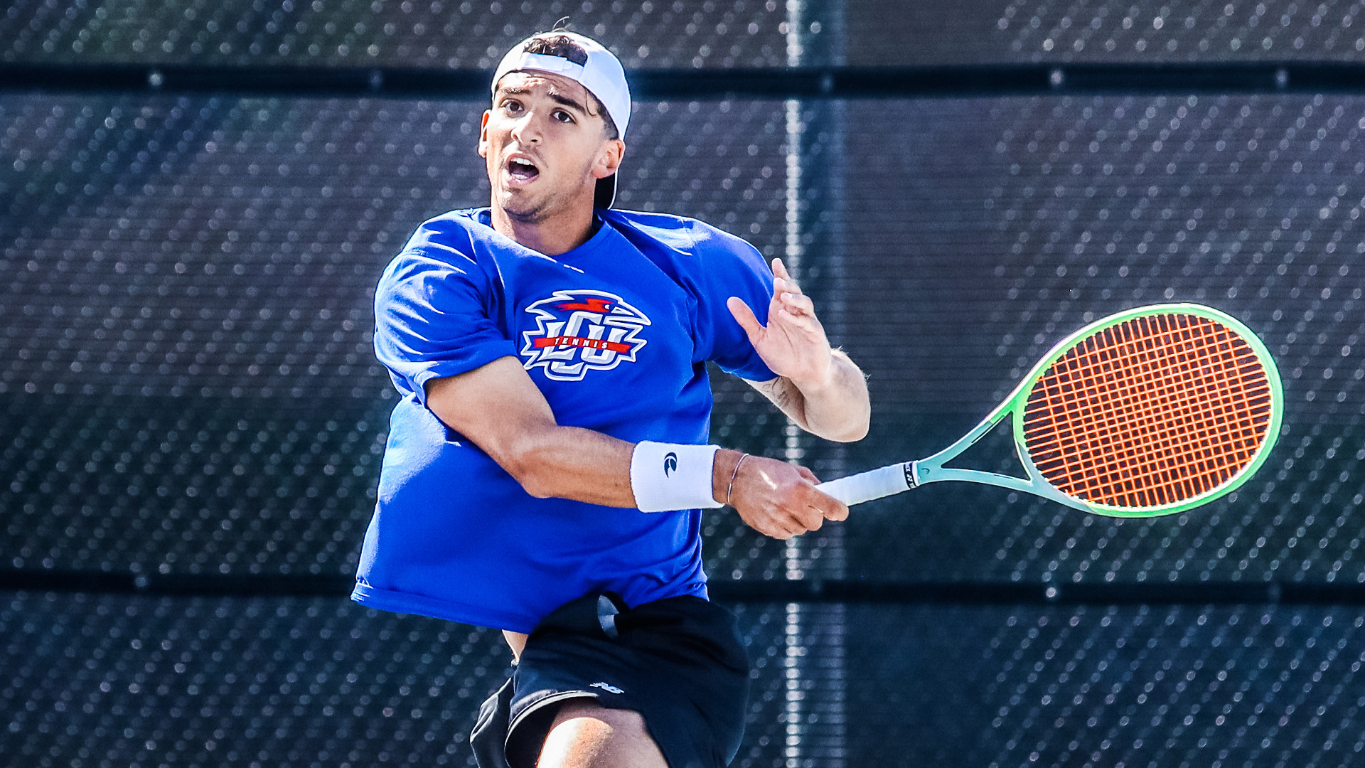 A Lubbock Christian University Chaps men's tennis player wearing a blue LCU t-shirt, black shorts, a white wristband, and a white backwards cap is captured mid-swing hitting a forehand shot with a green and orange racket. His mouth is open and his eyes are focused upward tracking the ball. A dark tennis court fence is visible in the background.