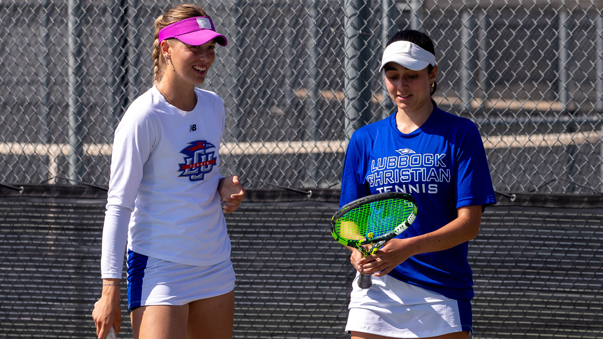 Two Lubbock Christian University Lady Chaps tennis players stand together on an outdoor tennis court in front of a chain-link fence. The player on the left wears a white New Balance long-sleeve shirt with the LCU Tennis logo and a bright pink visor, and is smiling. The player on the right wears a blue 