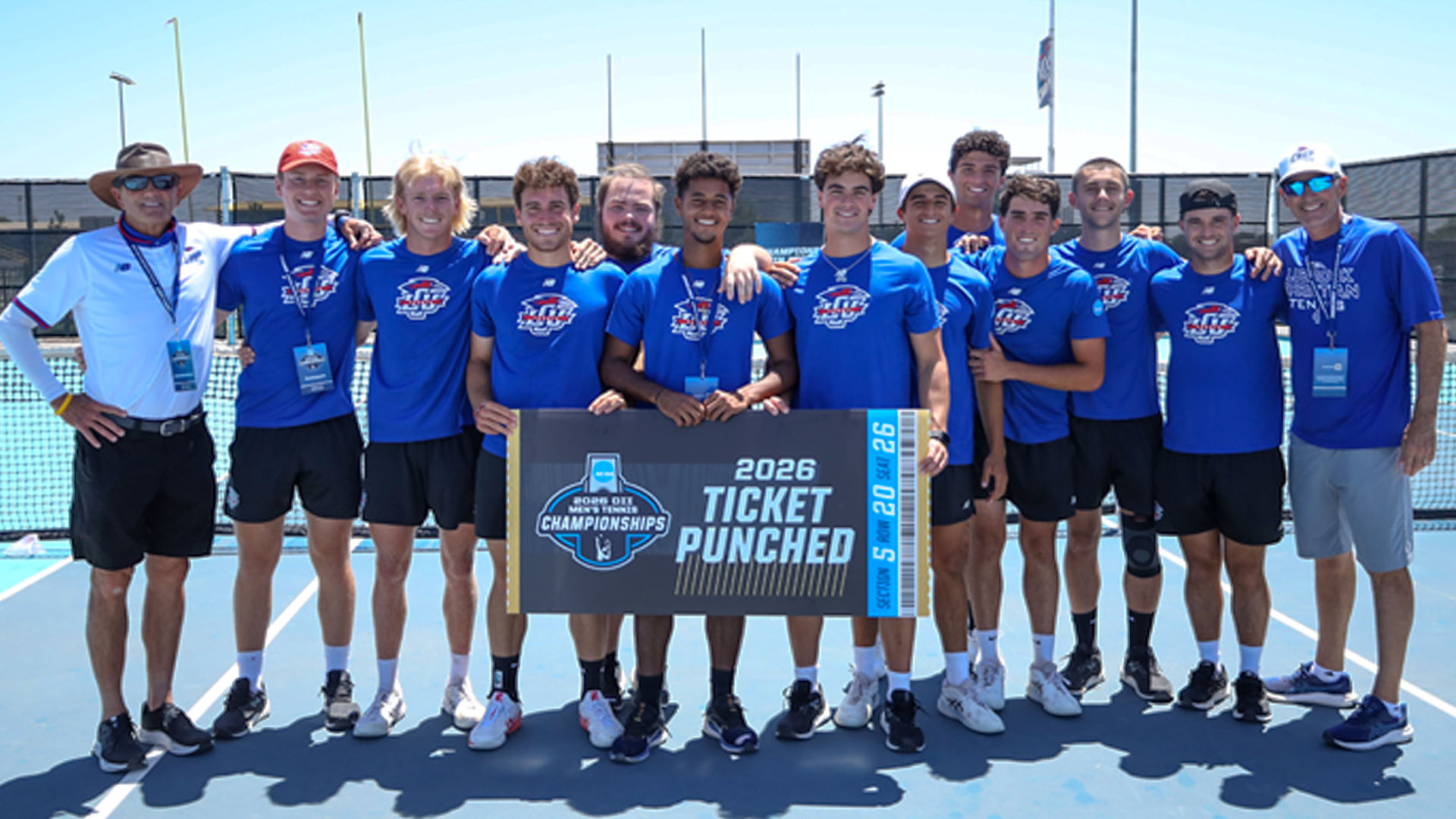 The Lubbock Christian University Chaps Men's Tennis team and coaching staff pose together on a sunny outdoor blue tennis court, all wearing blue LCU New Balance uniforms. The group of approximately thirteen players and coaches stands in a row with arms around each other, holding a large 