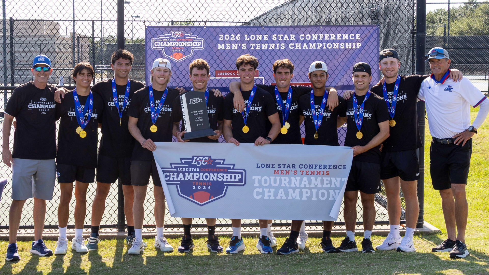 The Lubbock Christian University Chaps Men's Tennis team poses together outdoors on a sunny day in front of the 2026 Lone Star Conference Men's Tennis Championship backdrop. The group of approximately eleven players and two coaches stands on a grass surface, all wearing black LSC Tournament Champions t-shirts and gold medals on blue lanyards. A player in the center holds the championship trophy, and the group holds a large LSC Lone Star Conference Men's Tennis Tournament Champion 2026 banner in front of them. A chain-link fence and trees are visible in the background.