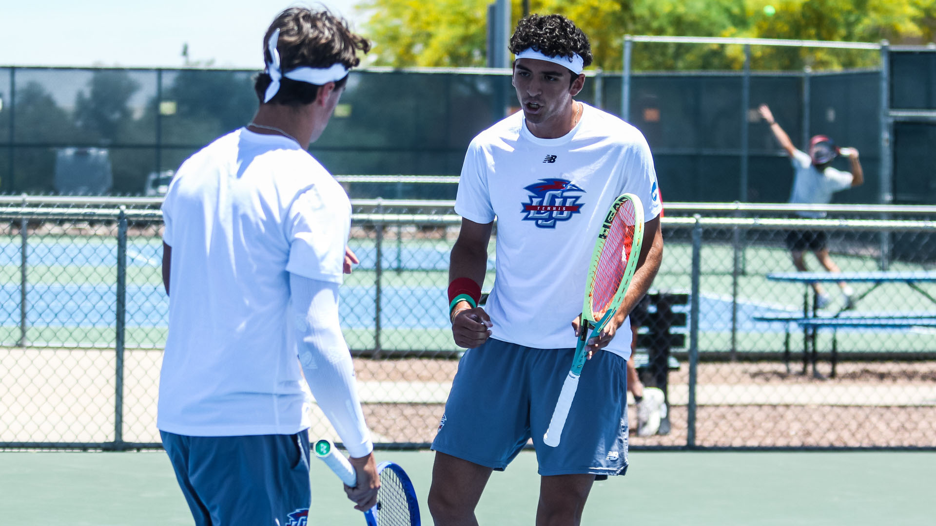 Action photograph taken during an LCU men's tennis doubles match on an outdoor hard court. In the foreground, two LCU players in white New Balance shirts with the LCU Tennis logo and blue shorts face each other in a between-point exchange. The player on the right, wearing a white headband and a red wristband, holds a green and orange tennis racket and appears to be talking to his doubles partner. The player on the left, seen from behind and wearing a striped visor, holds a blue racket. In the background, a third person — partially visible near the fence — raises their arm in celebration. Additional tennis courts, chain-link fencing, and trees are visible in the background under a clear blue sky