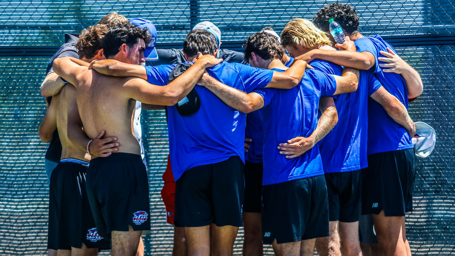 The LCU Chaparrals men's tennis team huddles together on an outdoor tennis court, arms around each other's shoulders and waists in a tight group embrace. Most players wear blue LCU shirts and black shorts, with LCU logos visible on several shorts. One player on the left is shirtless. A coach in a white shirt and blue cap is visible within the huddle. The group faces away from the camera toward a chain-link fence with a teal windscreen. The image conveys a moment of team unity, likely following the conclusion of a match