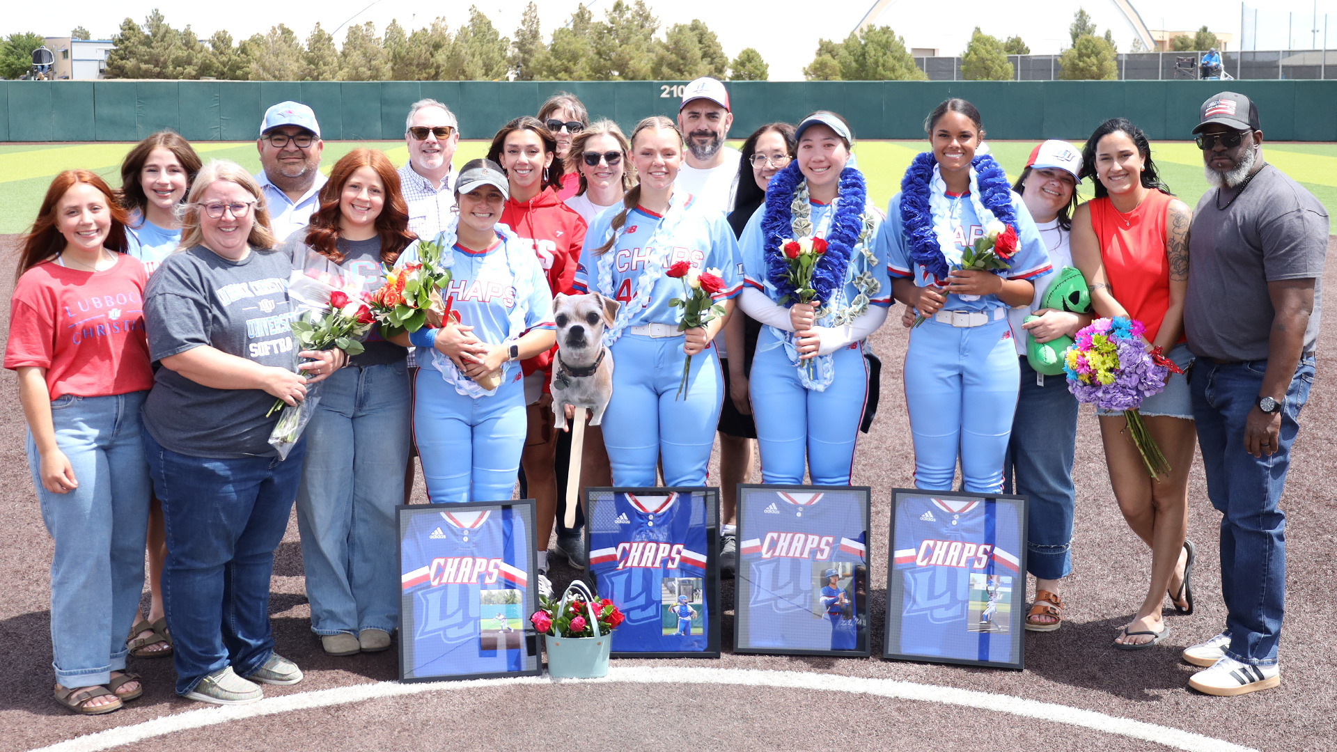A group photo on a softball field infield dirt circle during what appears to be a Senior Day celebration for the Lubbock Christian University Lady Chaps Softball team. Four players in light blue 