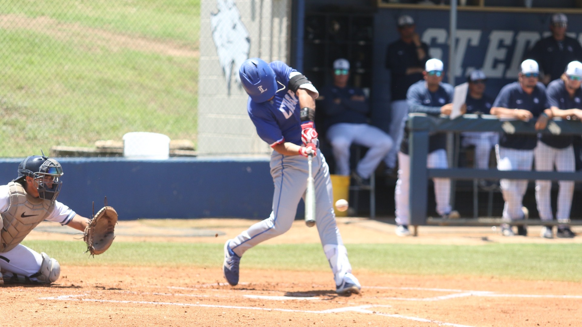This is a photo of Lubbock Christian catcher Ryan Gargett (blue jersey, red batting gloves, blue helmet) hitting a pitch for a home run during a game at Lucian Hamilton Field while members of the St. Edward's dugout look on.