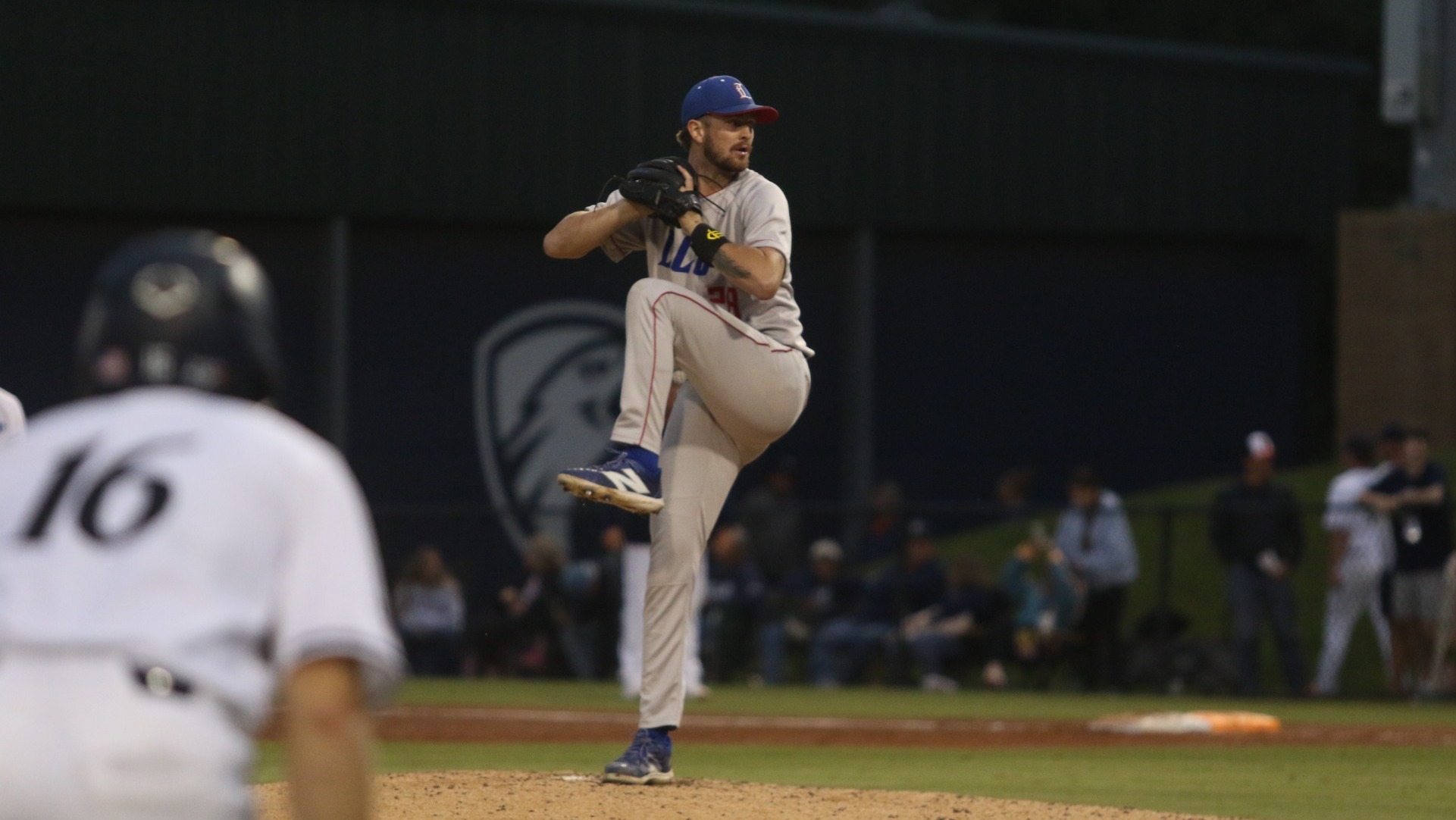This is a photograph of Lubbock Christian pitcher Caden Cline (grey jersey #28, grey pants, blue hat) winds up to throw a pitch during a game at UT Tyler's Irwin Field.