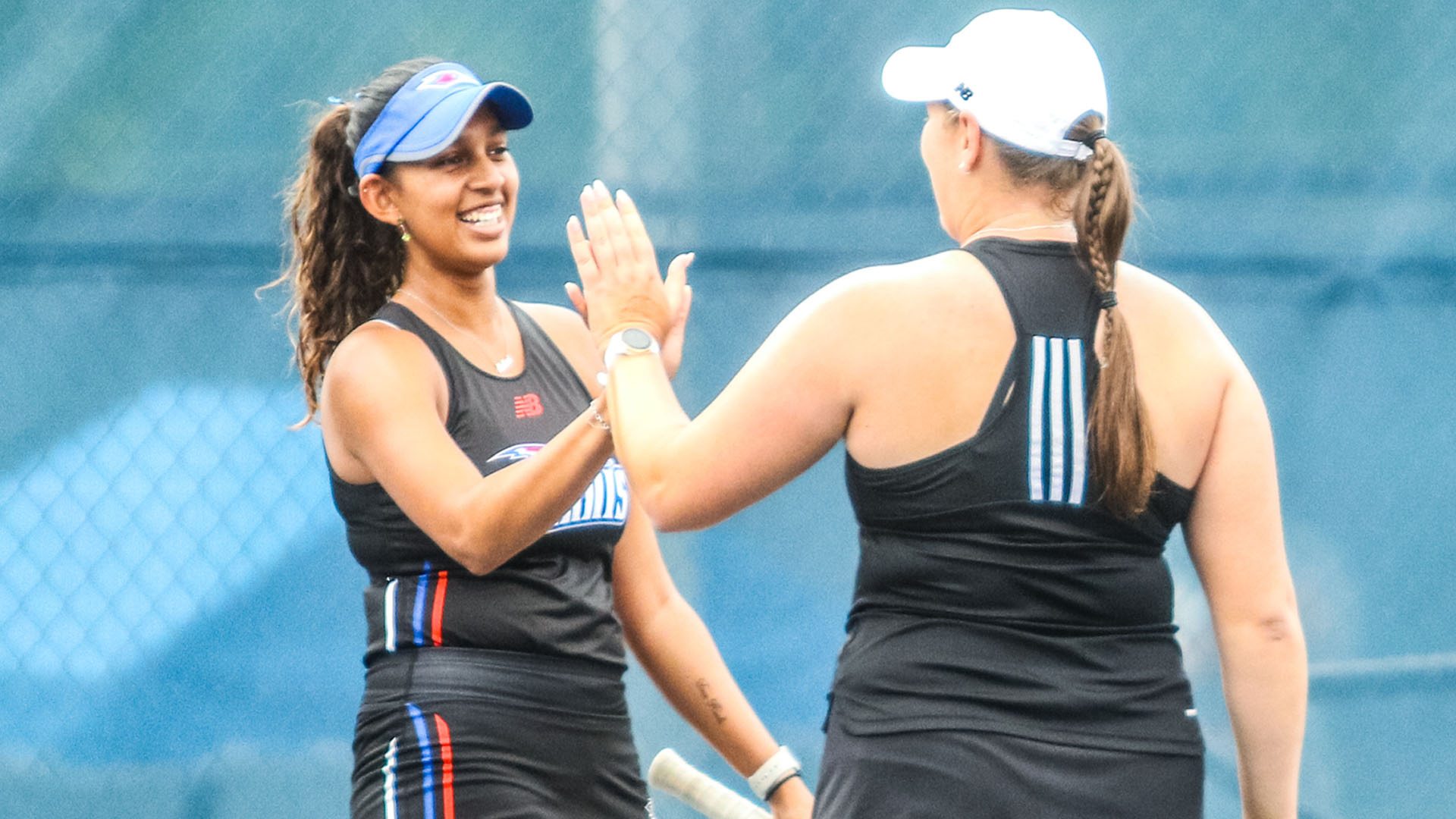 Two Lubbock Christian University Lady Chaps tennis players high-five each other on an outdoor tennis court during a doubles match. The player on the left, facing the camera, wears a black LCU Tennis New Balance uniform and blue visor and is smiling. The player on the right, shown from behind, wears a black tennis top with white stripes and a white New Balance cap with a braided ponytail, and holds a racket in her other hand. A blue chain-link fence is visible in the background.