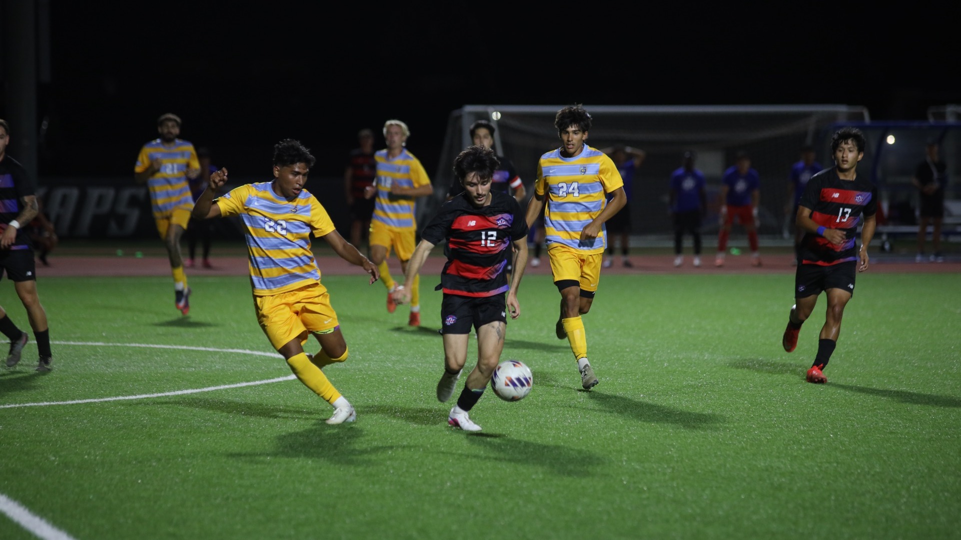 This is a photograph of Lubbock Christian men's soccer player Lucas Carli (black jersey, #12) dribbling the ball past several defenders from Fort Lewis (yellow jerseys) during a game at the LCU Soccer & Track Complex. 