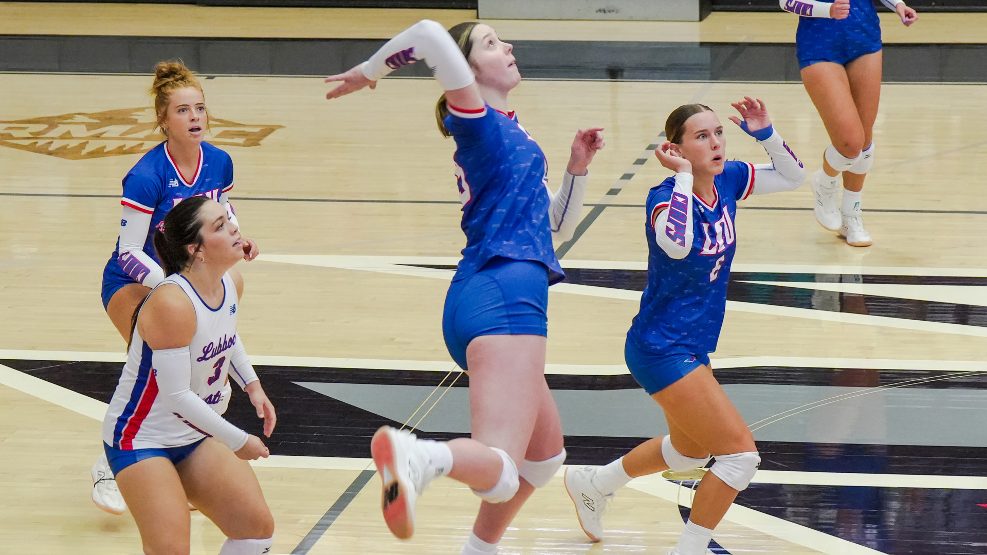 Four LCU volleyball players in blue and white Lubbock Christian New Balance uniforms track the ball during match action. A player in a blue LCU jersey leaps to attack at center court while number 6 in blue reads the play beside her. Number 3 in a white Lubbock Christian jersey and a teammate in blue crouch in ready position behind them. The action takes place on a light wood court with black and white floor markings visible.