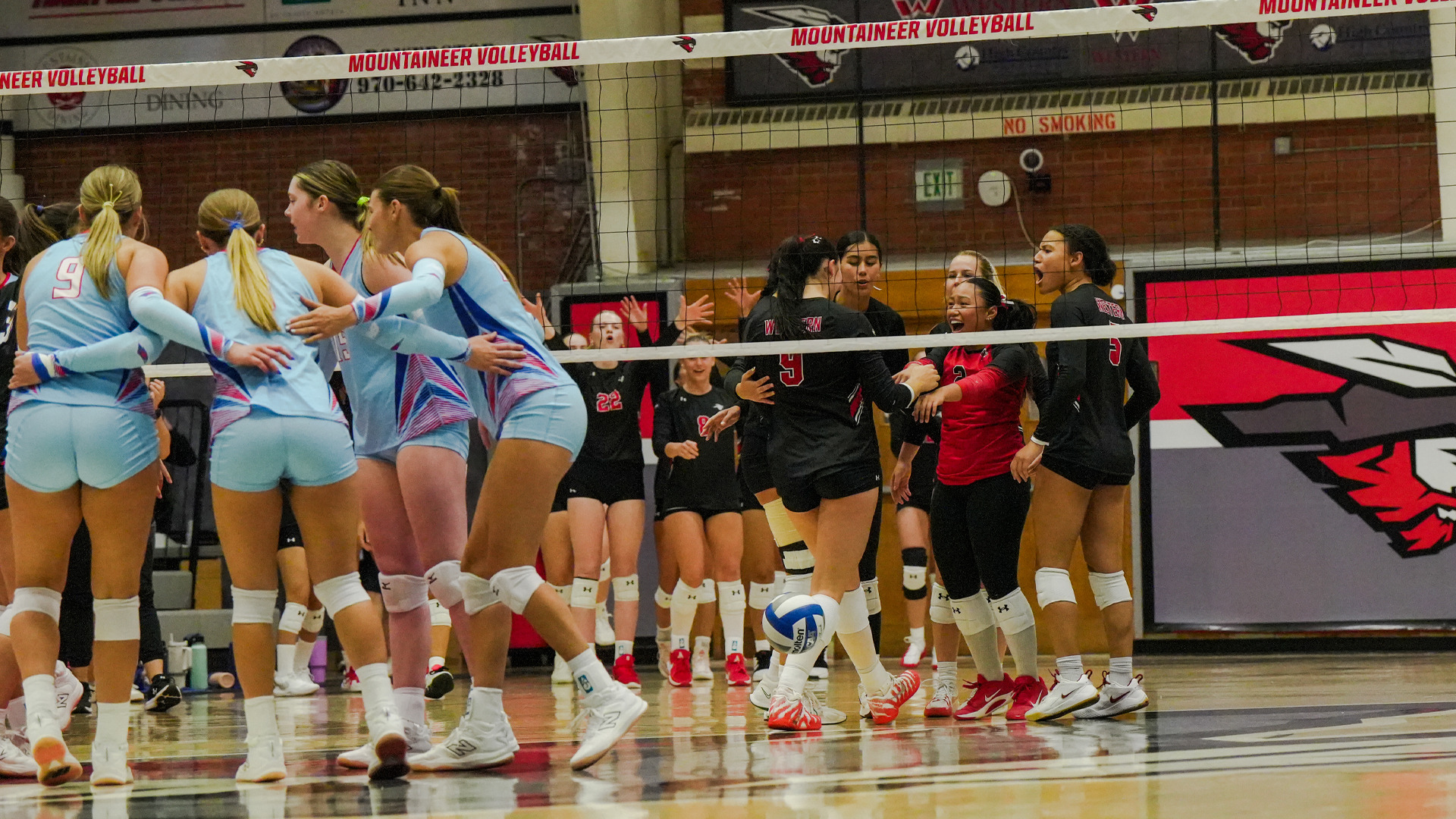 Both teams react after a point during the LCU vs. Western Colorado match at Paul Wright Gym. On the left, LCU players in light blue uniforms — including number 9 — huddle together after the play. On the right, Western Colorado Mountaineers players in black and red uniforms celebrate exuberantly near the net, with a volleyball rolling on the floor between the teams. The 