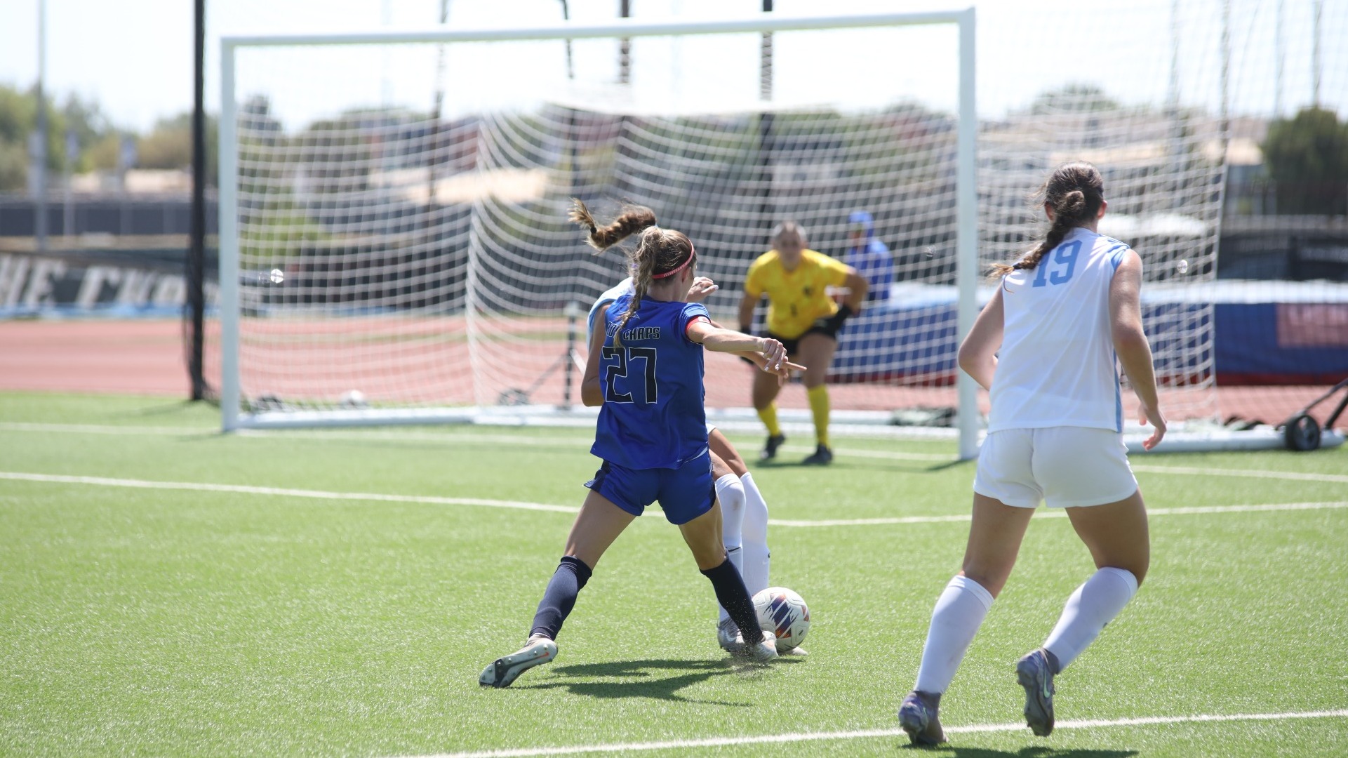 This is a photo of Lubbock Christian forward Claire Jacobson (blue jersey, #27) attempting to dribble between two Fort Lewis defenders (white jerseys) during a game at the LCU Soccer & Track Complex. 