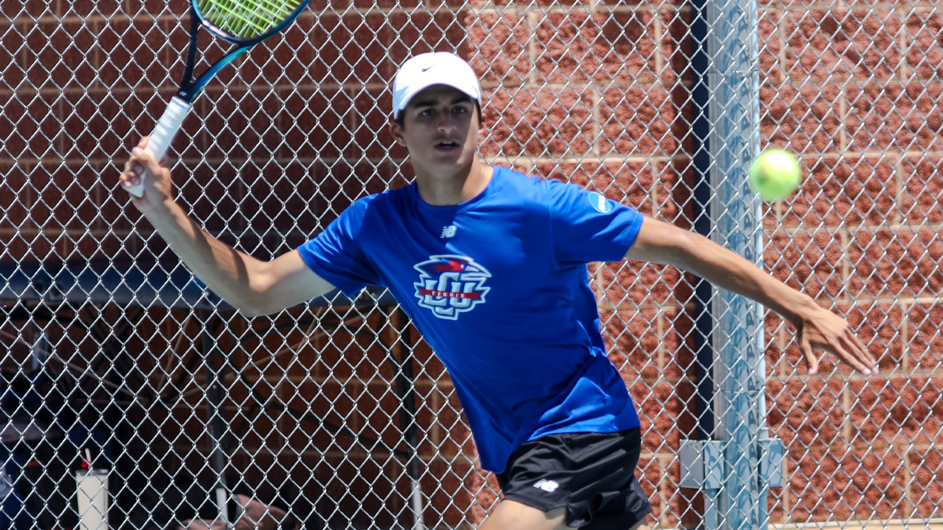 An LCU men's tennis player in a blue LCU Tennis New Balance shirt, black shorts, and white Nike cap reaches to hit a forehand, eyes tracking a yellow tennis ball, with a chain-link fence and brick wall visible in the background