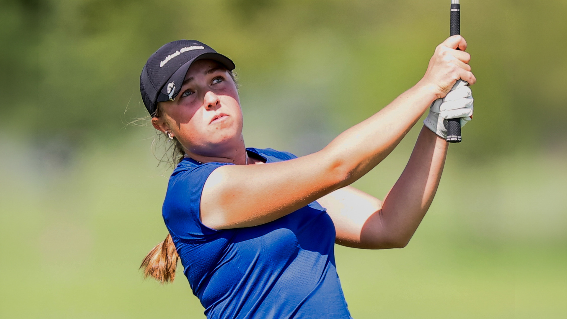 LCU women's golfer Abby Archunde follows through on a swing, eyes tracking the ball upward, wearing a blue polo and black Callaway cap with her auburn ponytail trailing behind her, against a blurred green fairway background