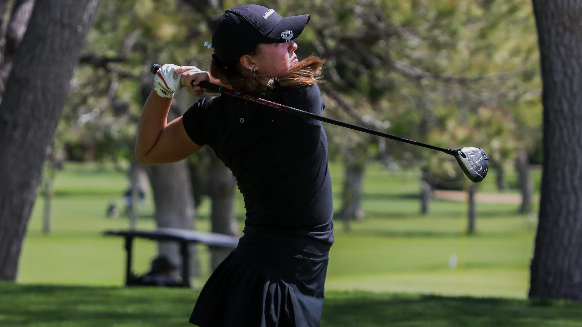 An LCU women's golfer in a black polo, black skirt, and black Callaway cap follows through on a driver swing, her ponytail trailing behind her, with tree-lined fairway and green grass visible in the background