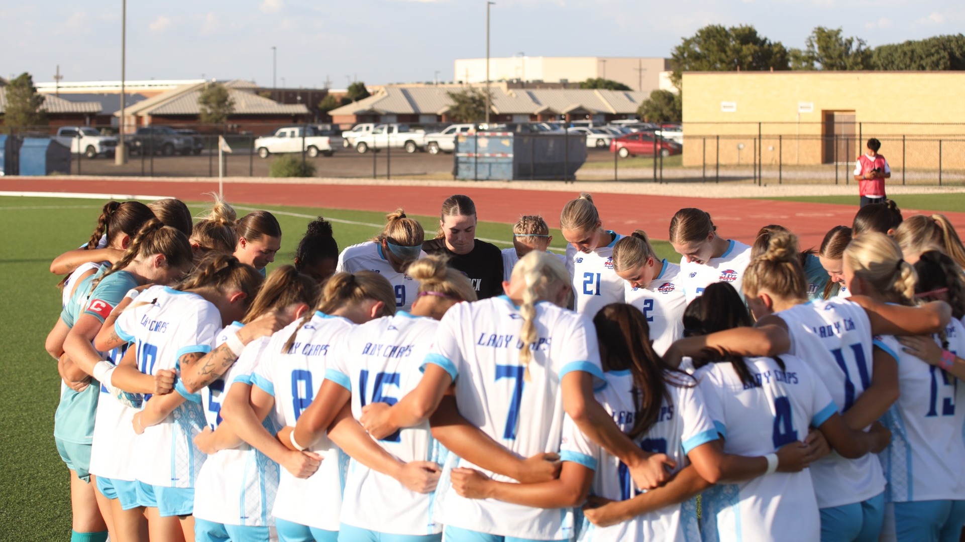 This is a photo of the Lubbock Christian women's soccer team, wearing white jersey, huddled up and praying before a match at the LCU Soccer & Track Complex. 