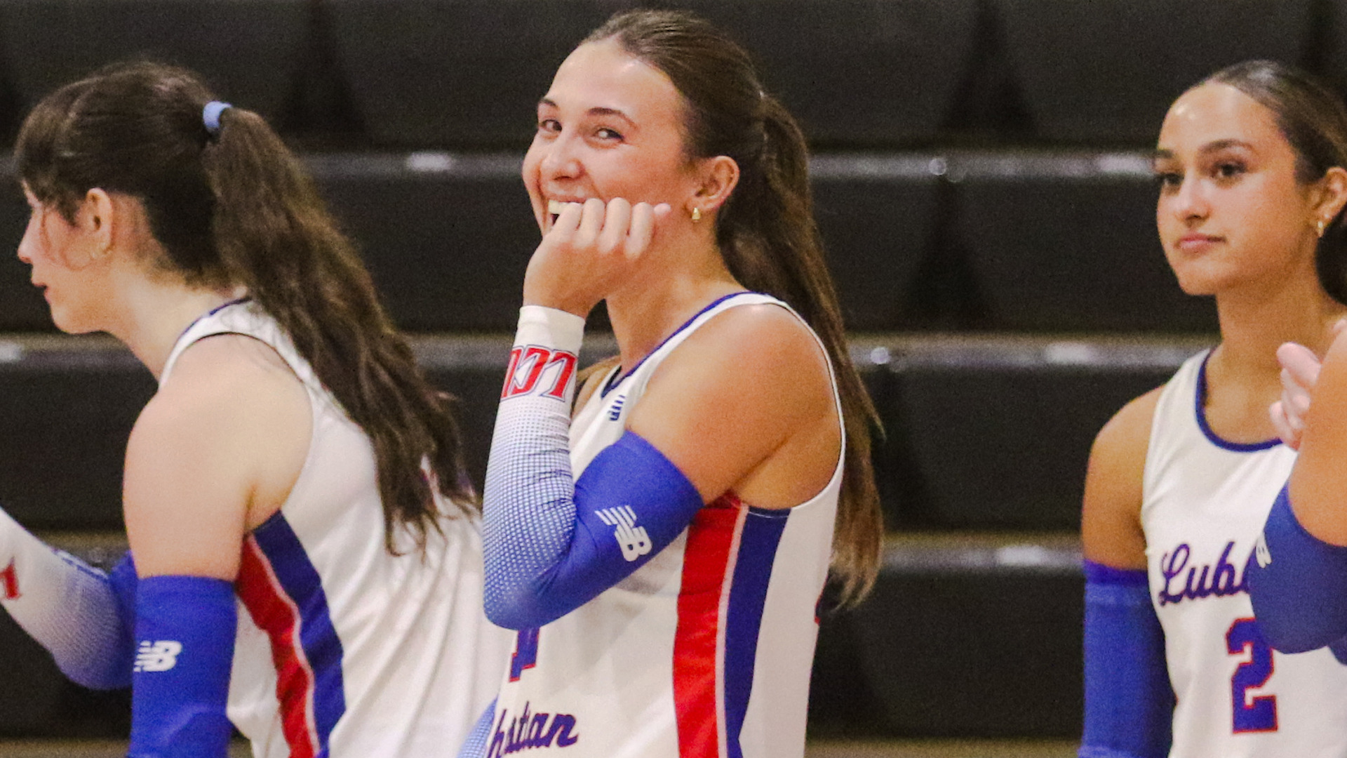 Close-up action photo of three volleyball players in white LCU jerseys with red and blue trim, standing in a row on the court in front of dark empty bleachers. The center player, with a ponytail, smiles broadly with her hand near her chin while looking off to the side. A player on the left is shown in profile, and a player on the right with dark hair is visible, her jersey reading 