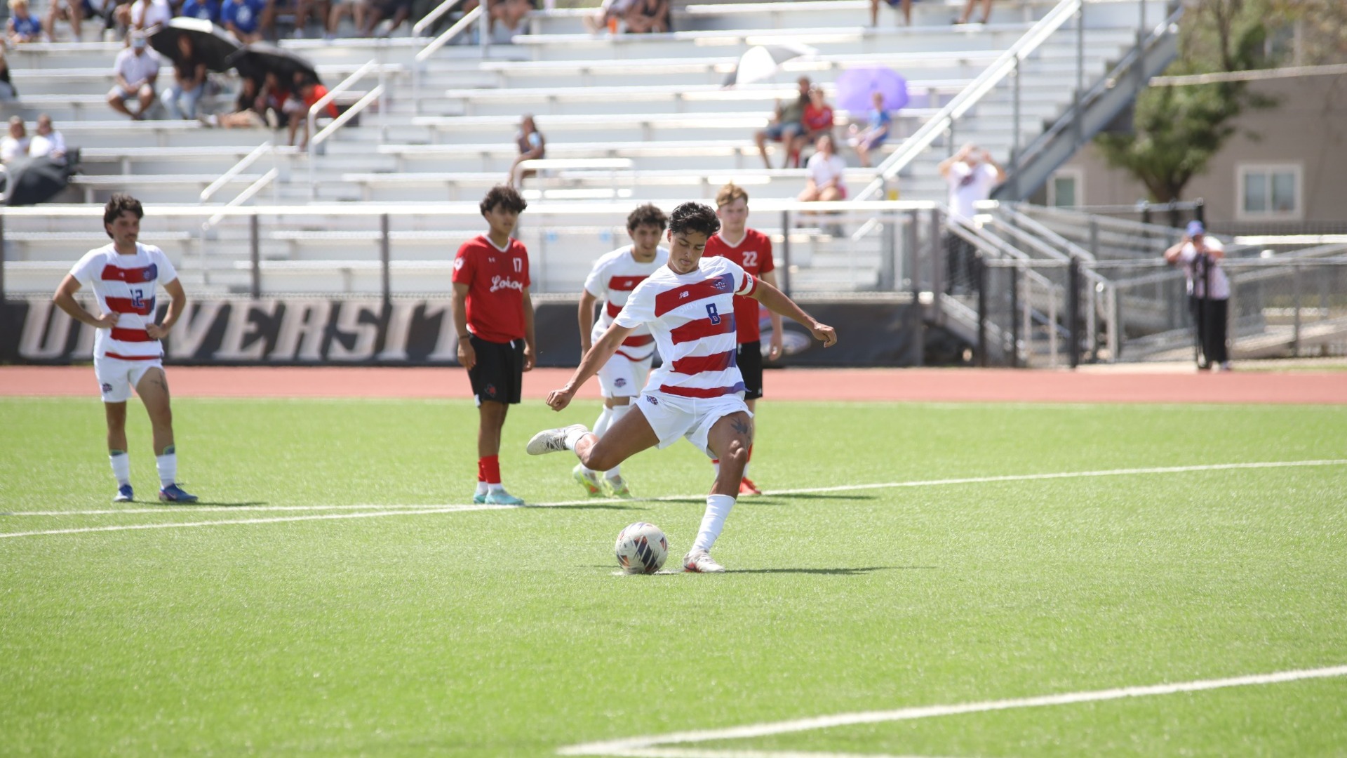 This is a photo of Lubbock Christian midfielder Aron Depaula (white jersey, #8) taking a penalty kick while Sul Ross State players (red jerseys) look on during a game at the LCU Soccer & Track Complex. 