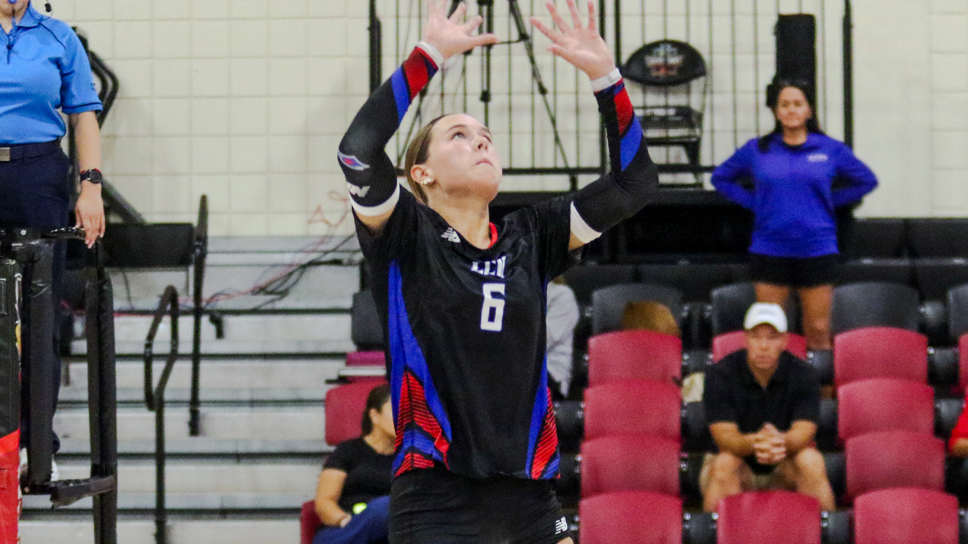 Action photo of LCU player #6, in a black jersey, setting the ball with both hands raised overhead, eyes tracking upward. A referee stands at left and a coach in a purple shirt stands at right, with red gym bleachers and seated spectators in the background.