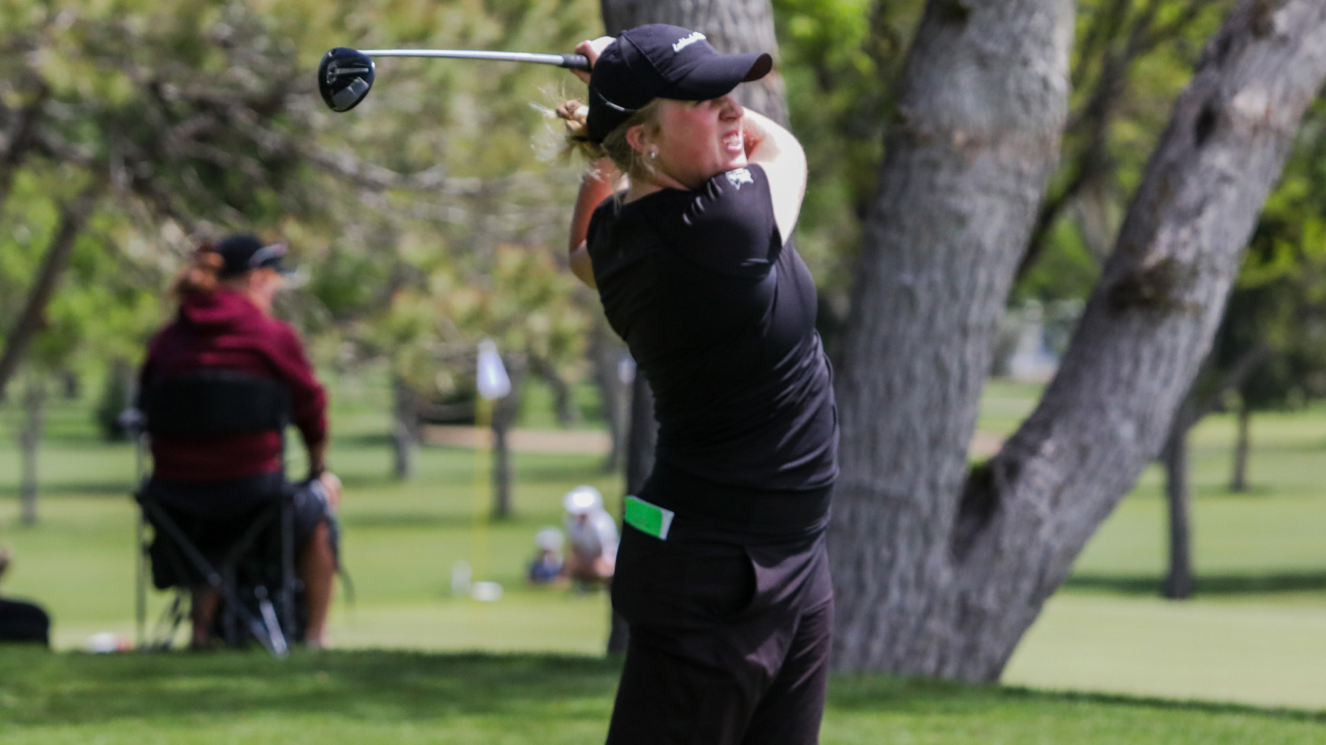 Action photo of a golfer in a black cap and black outfit completing her swing, club raised behind her, watching the ball's flight. She's on a tree-lined course, with a spectator seated in a folding chair in the background and a flag visible on the green in the distance.