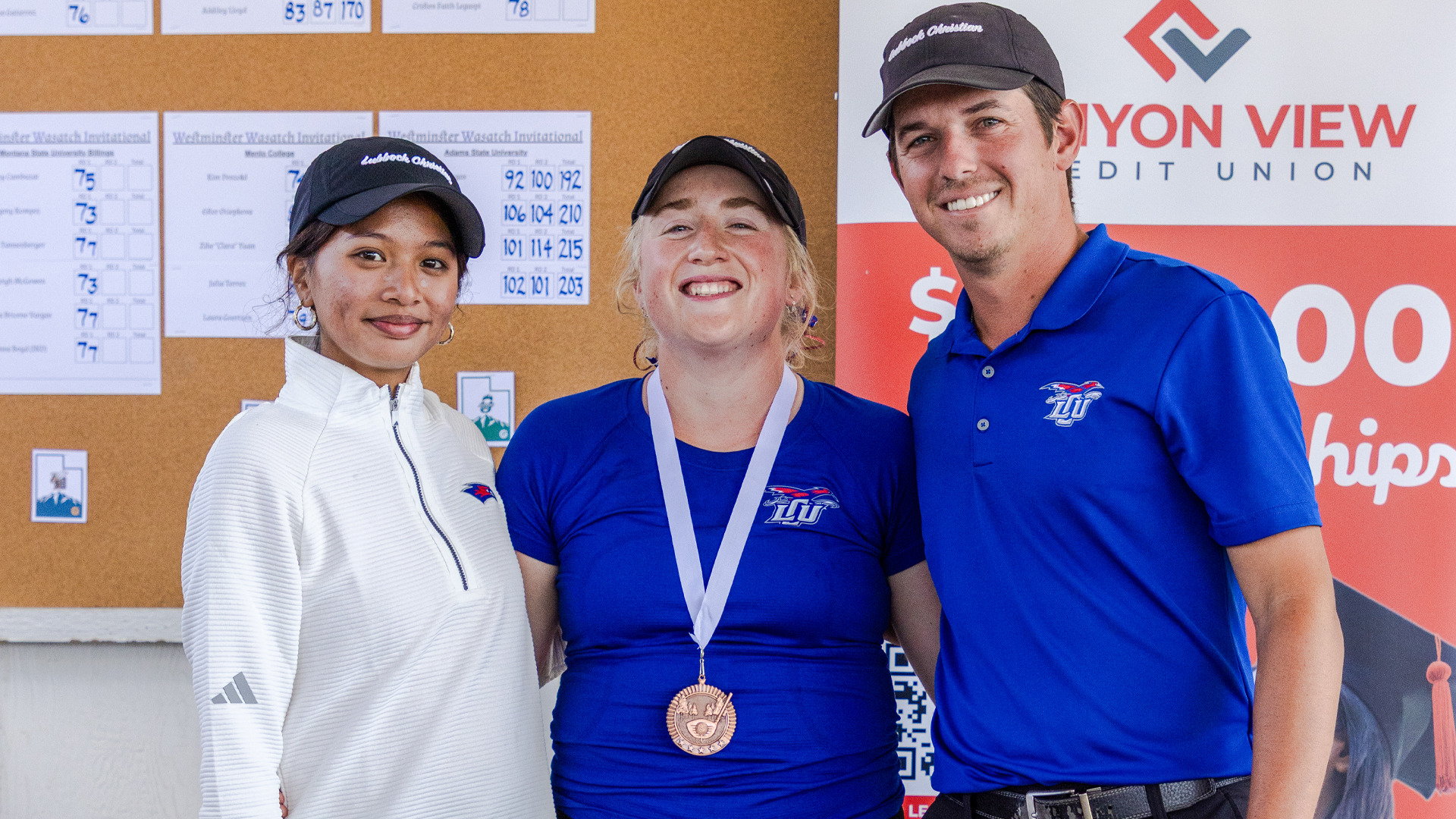 Three people pose smiling for a photo indoors near a scoreboard corkboard. A woman in a white quarter-zip stands at left, a woman in a blue LCU shirt wearing a bronze medal stands in the center, and a man in a blue LCU polo and dark cap stands at right with his arm around her. A 