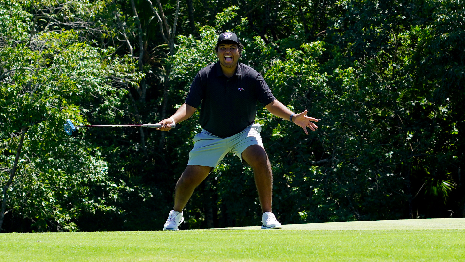 Man in a dark polo and gray shorts reacts excitedly on a putting green, crouching with a putter in one hand and the other hand gesturing, mouth open. Dense green trees fill the background under a clear blue sky.