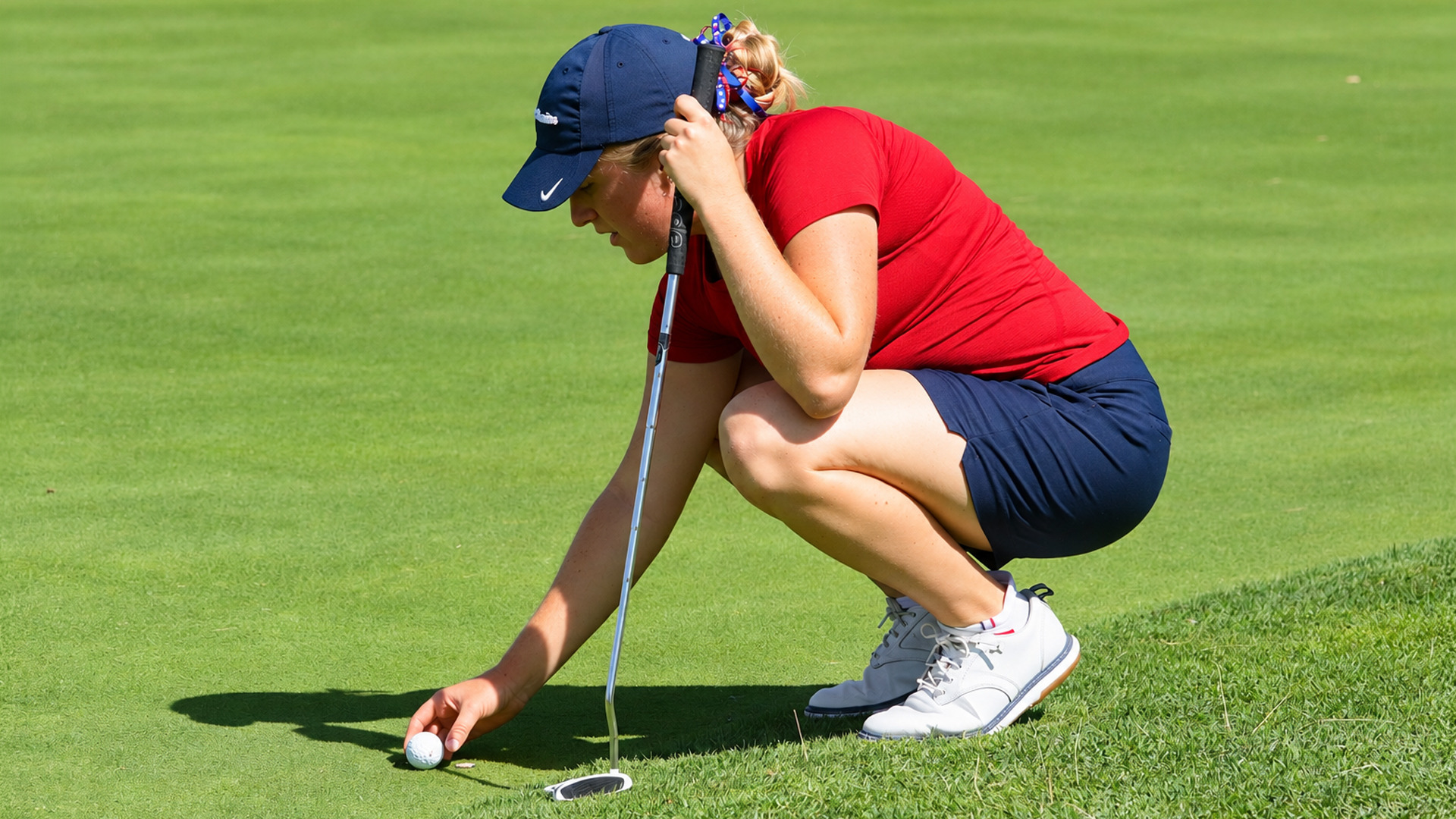 Golfer in a red shirt and navy shorts crouches on the green, placing or marking her ball with one hand while resting the other on her putter.