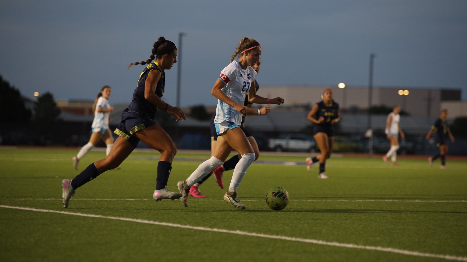 This is a photo of Lubbock Christian women's soccer player Claire Jacobson (white jersey, #27) dribbling the ball past two Wayland Baptist defenders (navy blue jerseys) during a game at the LCU Soccer & Track Complex. 