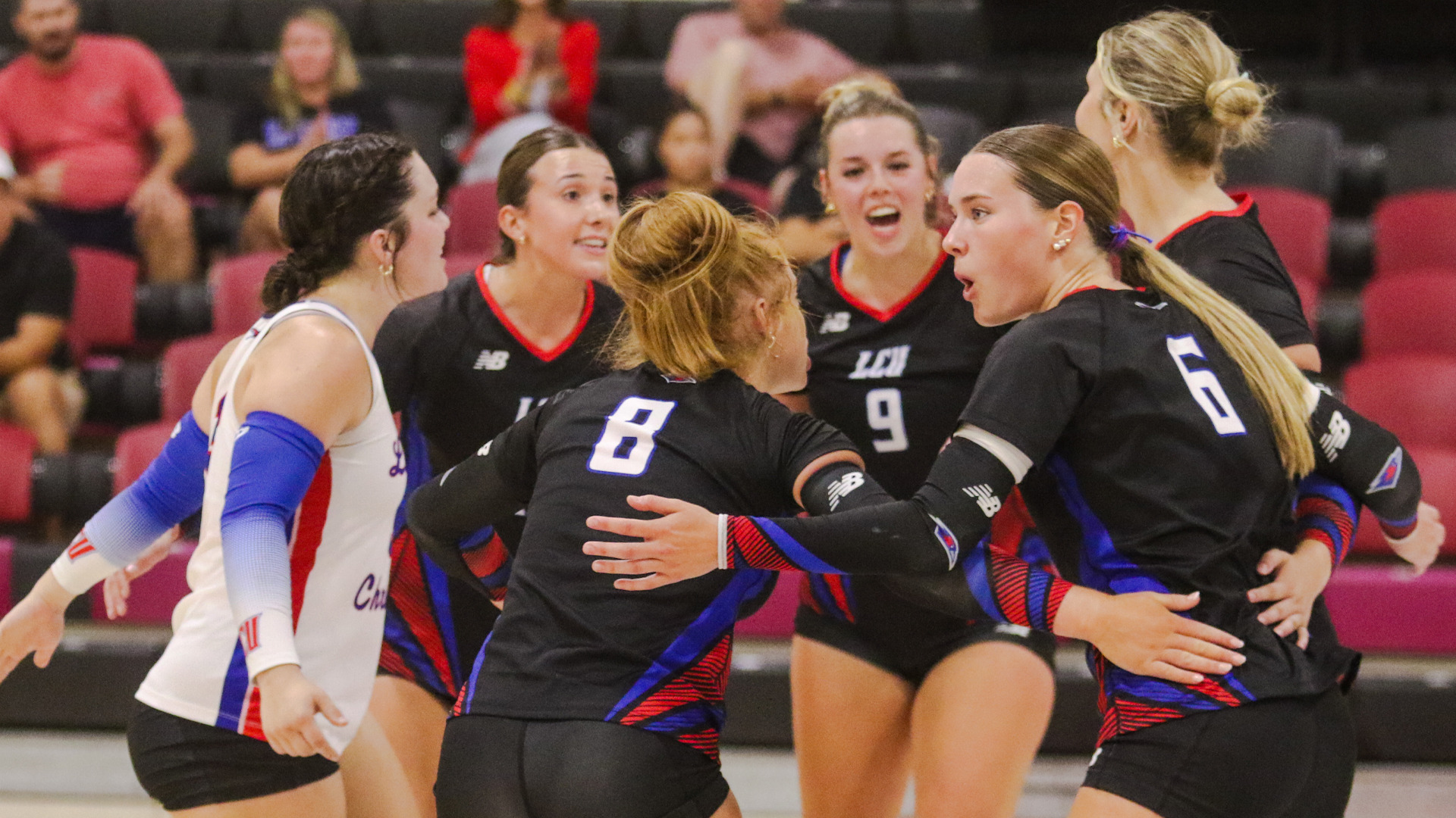 LCU volleyball players in black and red New Balance uniforms huddle together on the court after a point, with players number 8, 9, and 6 visible among the group reacting with intensity and excitement. A player in a white Lubbock Christian uniform with blue arm sleeves is visible on the left. Red bleachers with spectators are visible in the background at Barry University's Health & Sports Center in Miami Shores, Florida.
