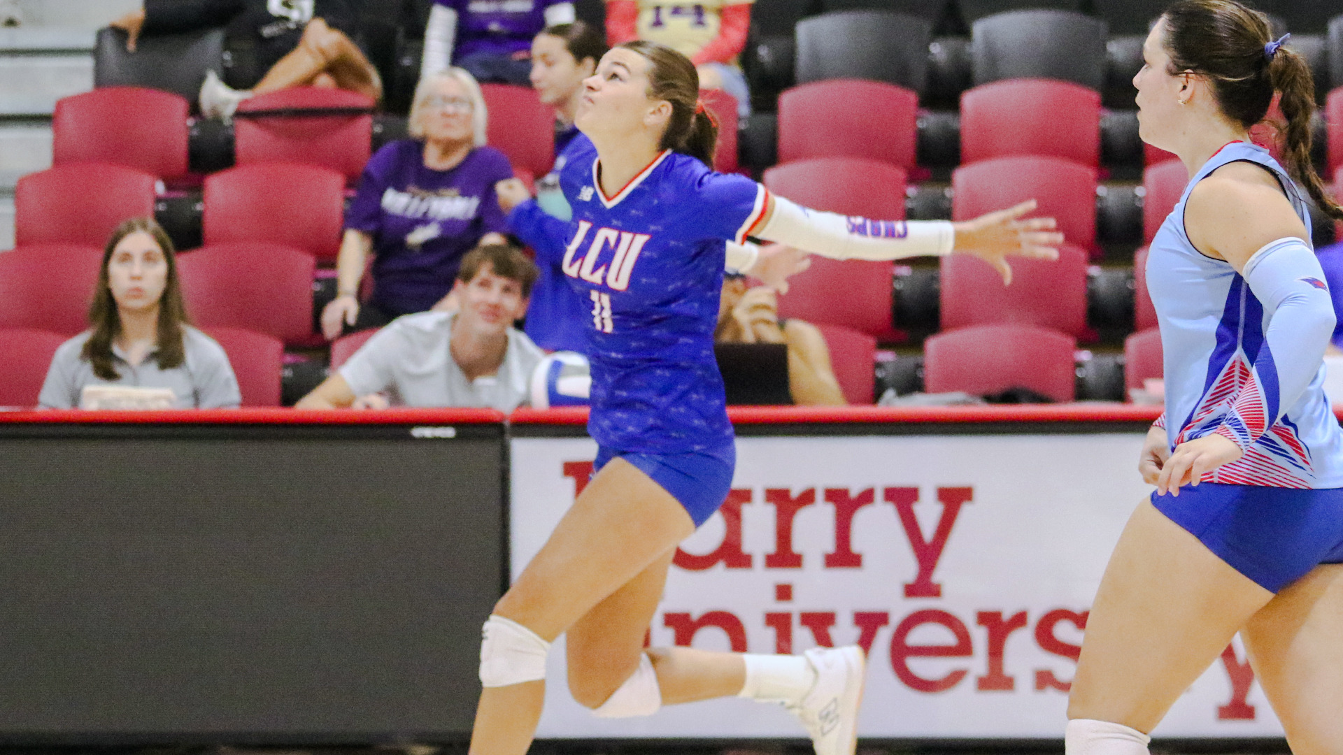An LCU volleyball player in a blue LCU number 33 jersey leaps to attack the ball, eyes locked upward with arms extended, while a teammate in a light blue uniform watches from the right. The 