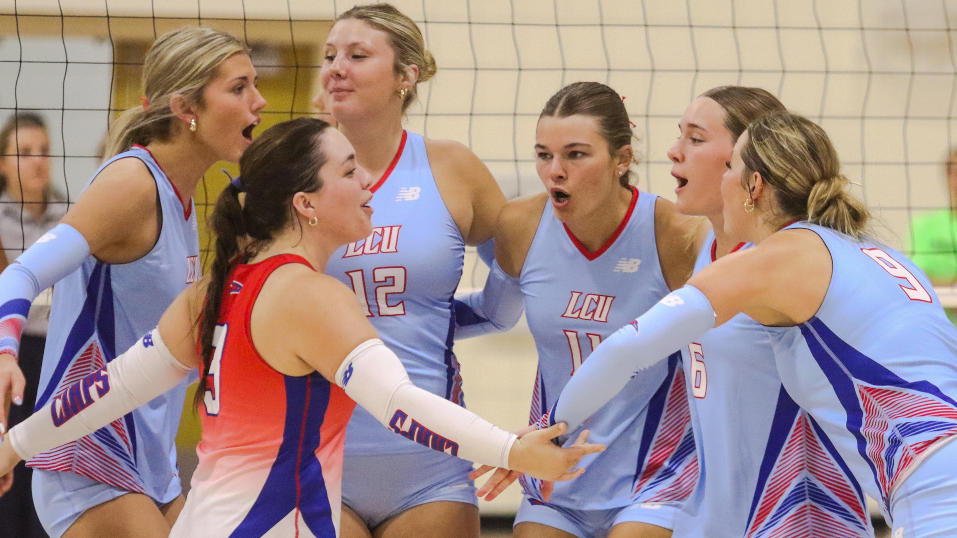 Six LCU volleyball players in light blue New Balance uniforms huddle together near the net during a match at Barry University, reacting with open mouths and intense expressions after a play. Players visible include numbers 12, 11, 6, and 9 in light blue LCU jerseys, with a player in a red and blue Chaps uniform at center. The volleyball net and a blurred background of spectators are visible behind them.