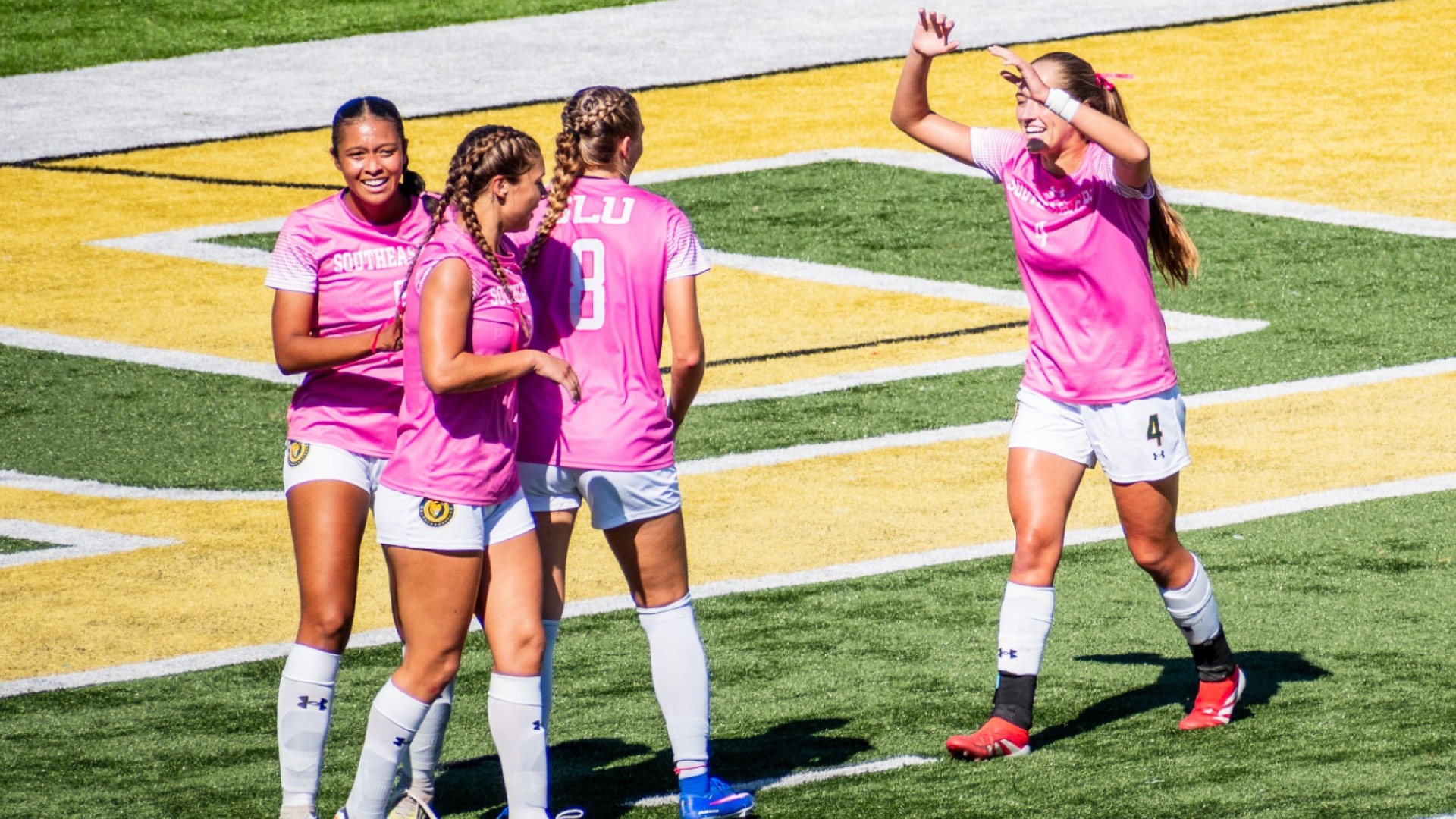 Multiple teammates in pink jerseys huddle and celebrate goal scored by Brooke Opferman