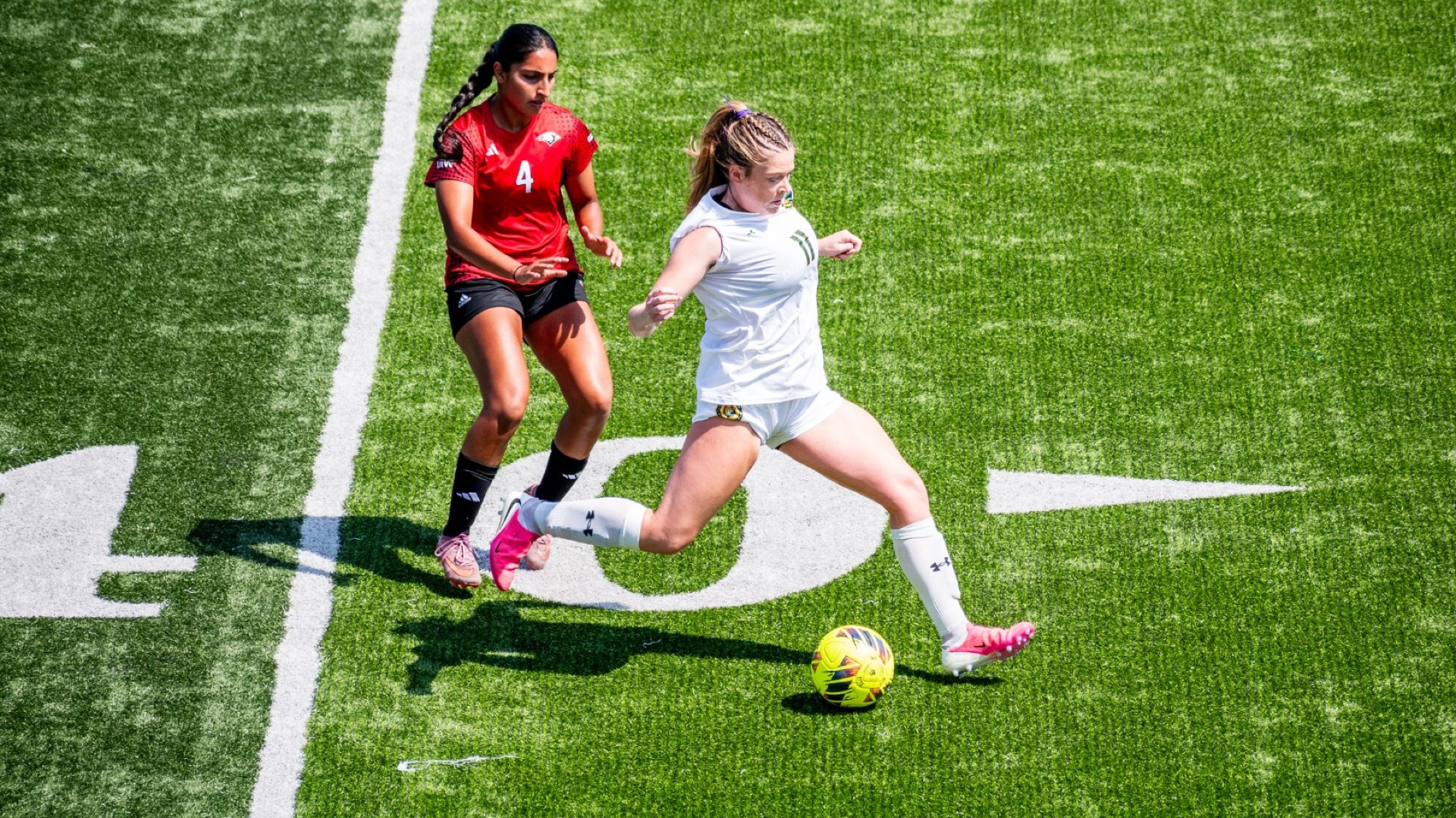 Defender Lainey Connell (white jersey) kicking the ball up the field and out of play vs UIW defender (red jersey)