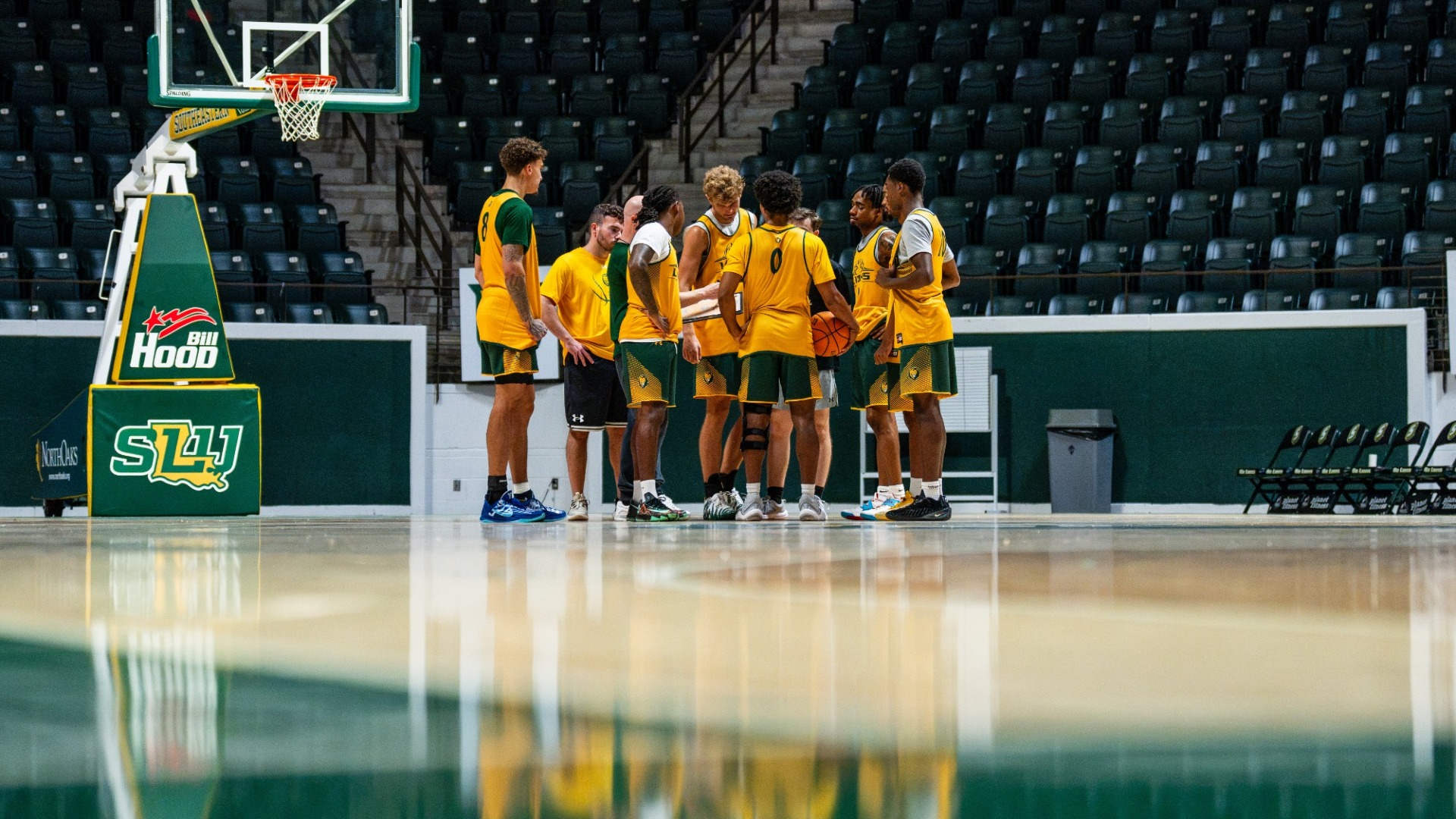 Several men's basketball players in a team huddle during practice alongside head coach and assistant coach