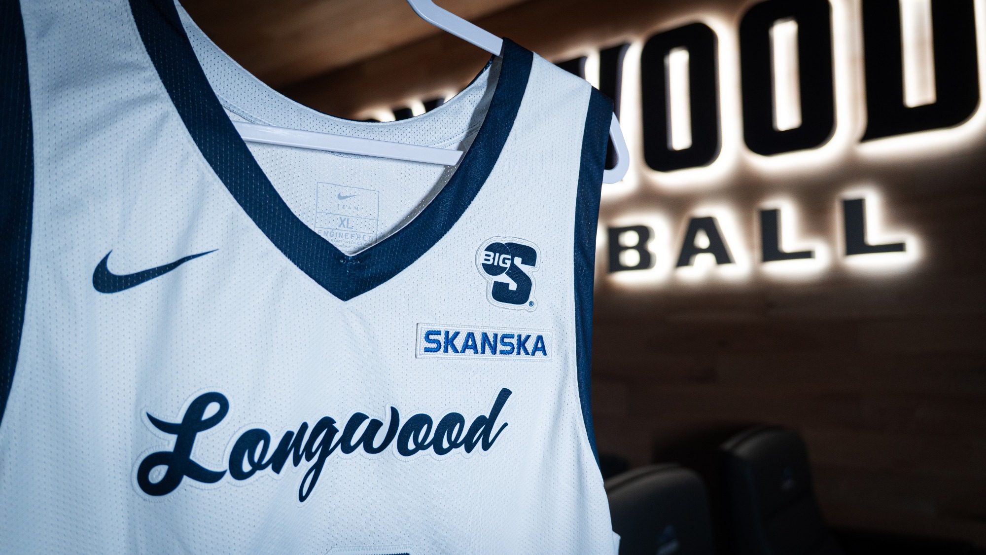 White Longwood basketball jersey with a Skanska patch on it and the Longwood Basketball lit on the wall behind it in the video room.