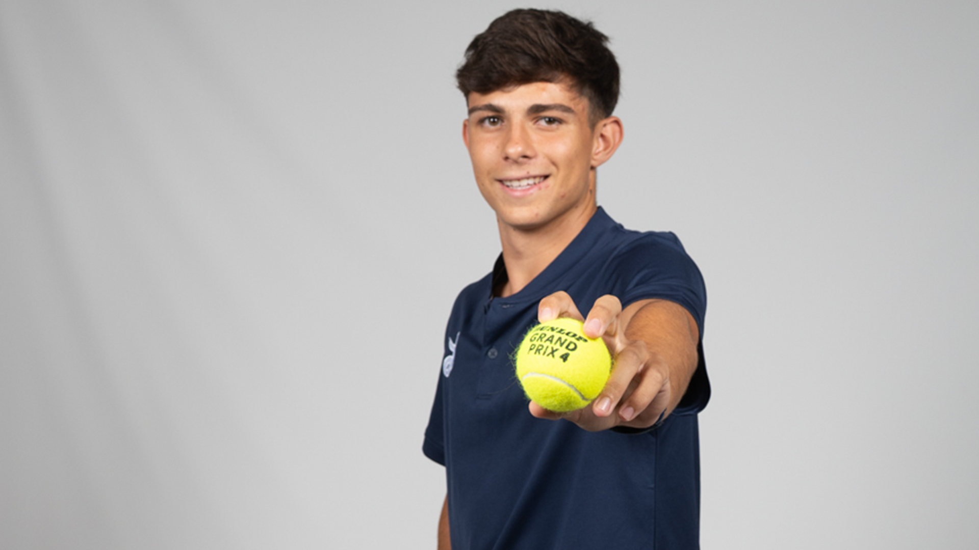 Marc Ferrrando poses in front of a gray backdrop holding a tennis ball towards the camera