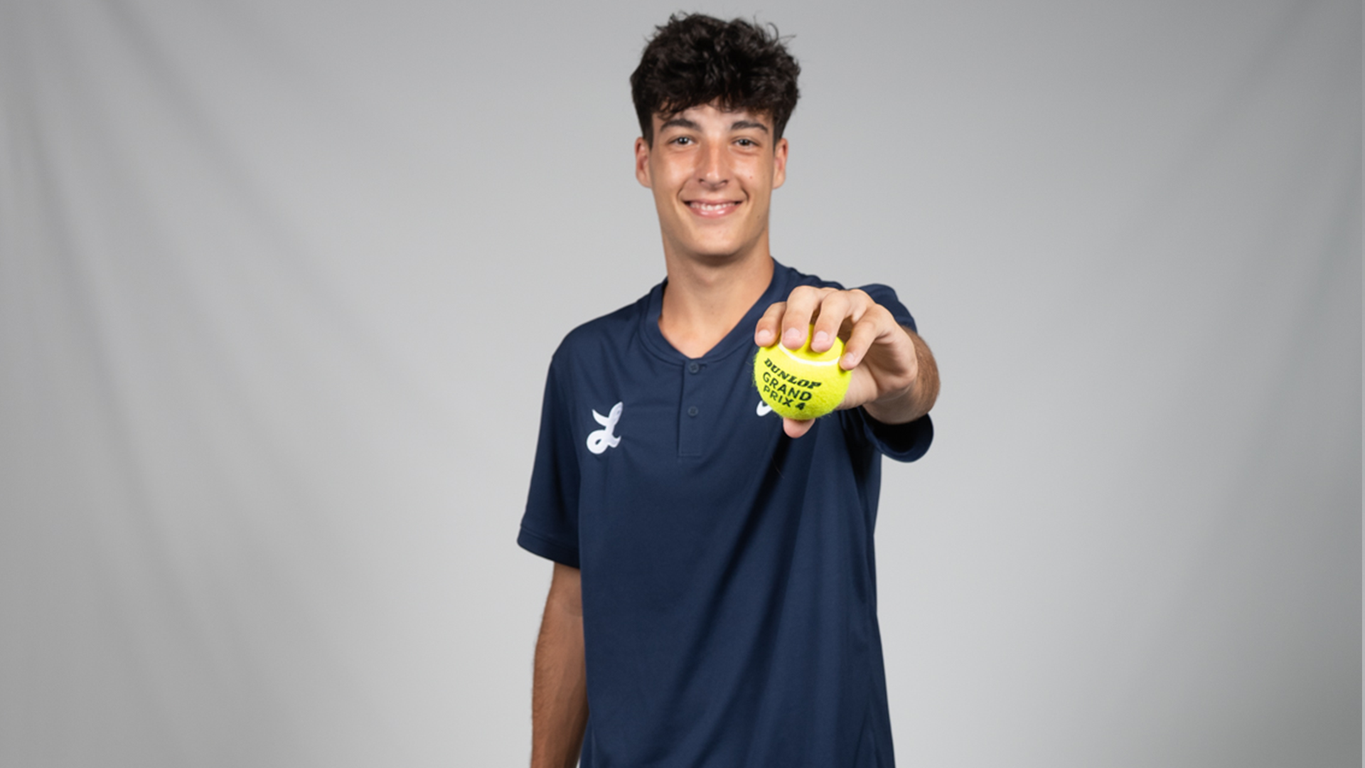 Rodrigo Guijarro poses in front of a gray backdrop holding a tennis ball to the camera