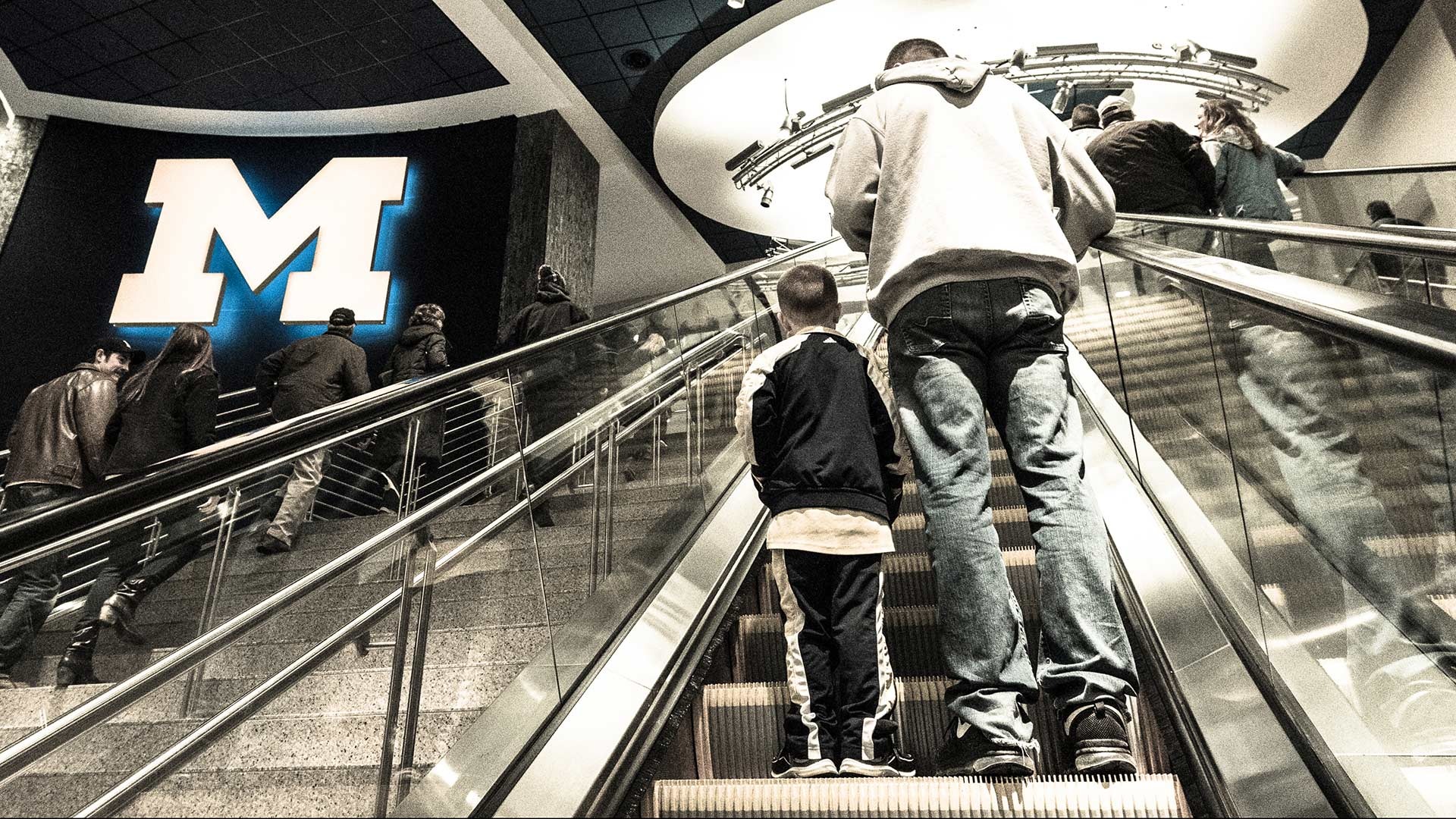 Fans riding the escalators inside Crisler Center near a large Michigan block M