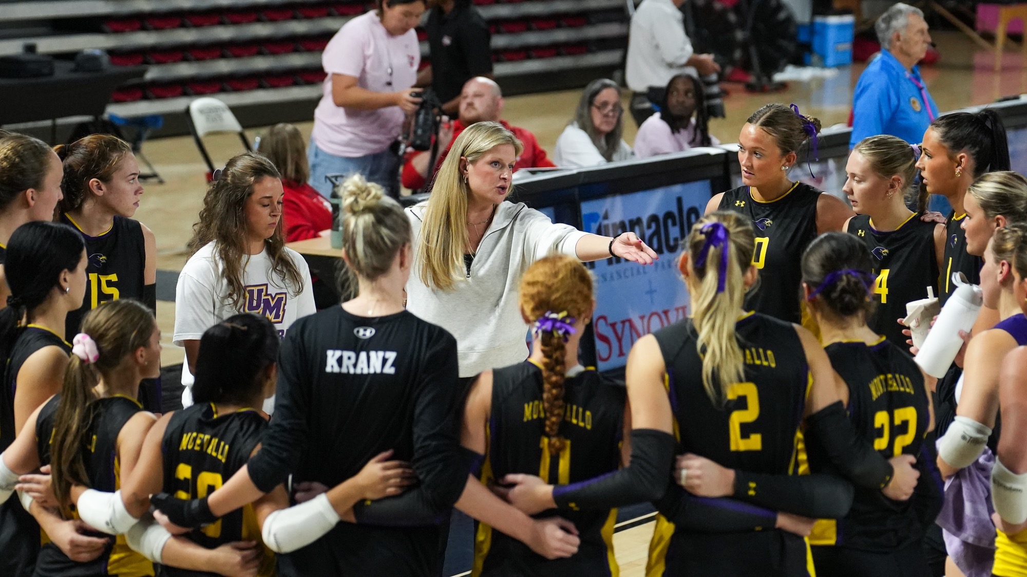Montevallo head volleyball coach Haven O'Quinn speaks to Falcons players gathered in a huddle during their match against Valdosta State. Players in black and gold Montevallo uniforms stand with their arms around one another while listening to their coach.