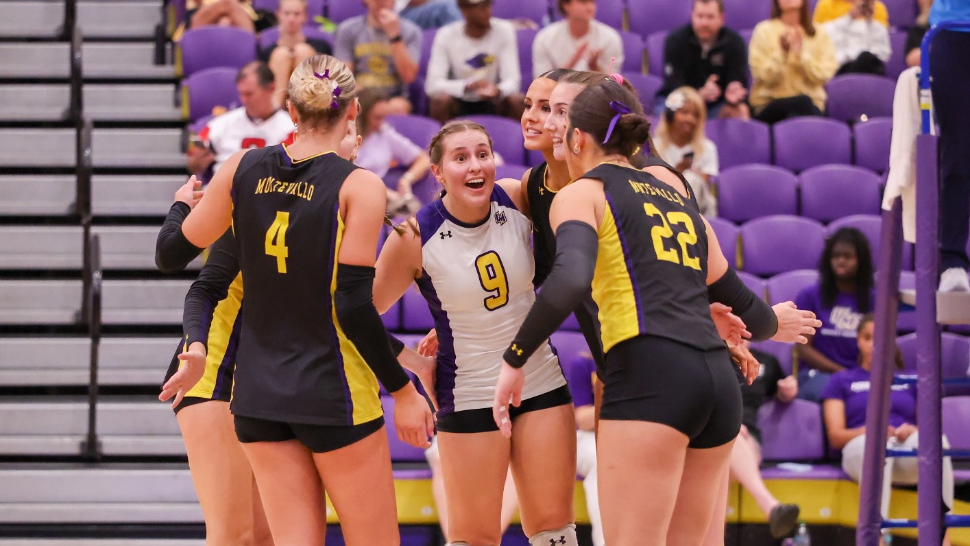 Montevallo volleyball players celebrate together on the court during their match against Spring Hill College. Players wearing black and gold jerseys, including No. 4 Ellie Scharlow and No. 22 Eliza Tyrrell, surround libero No. 9 Ava Wind in a white jersey, with spectators seated in purple bleachers in the background.