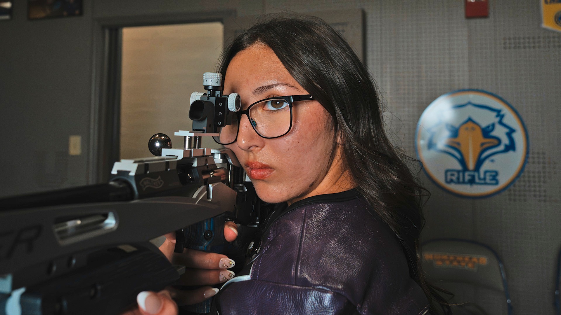 Dark-haired girl wearing a black top and clear shooting glasses aiming an air rifle