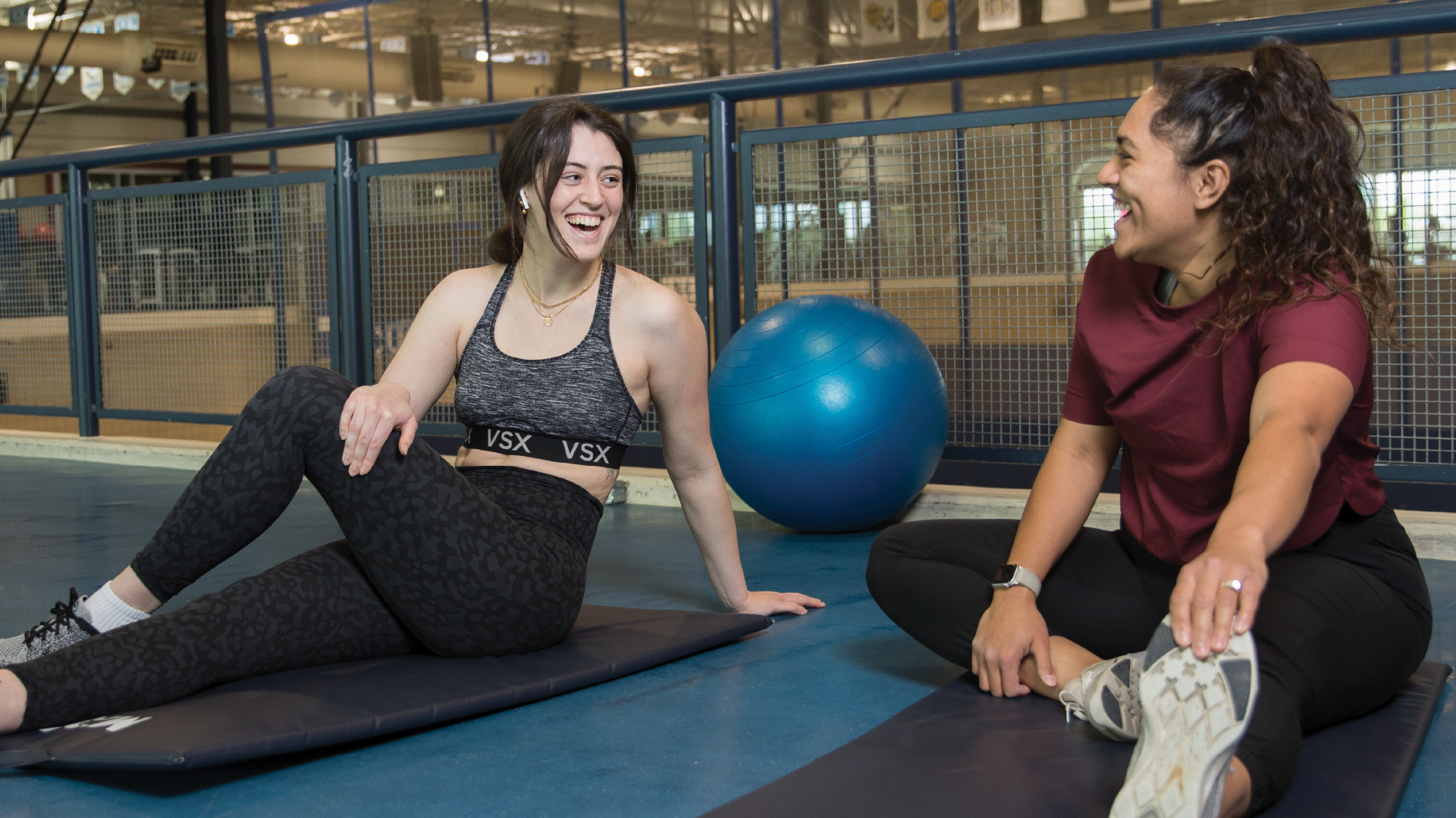 Two women in athletic wear smile and chat while stretching on yoga mats in an indoor gym, with a blue exercise ball in the background.