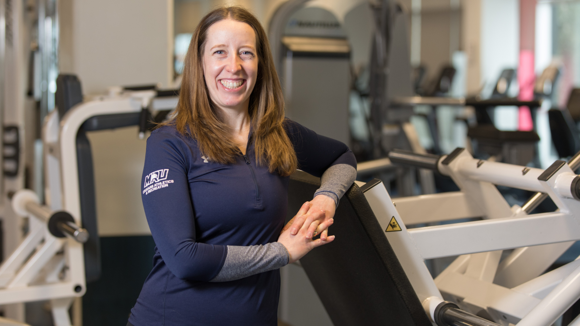 Sarah Hall, certified MRU personal trainer, smiles and leans against a piece of equipment in the fitness area with her hands clasped.