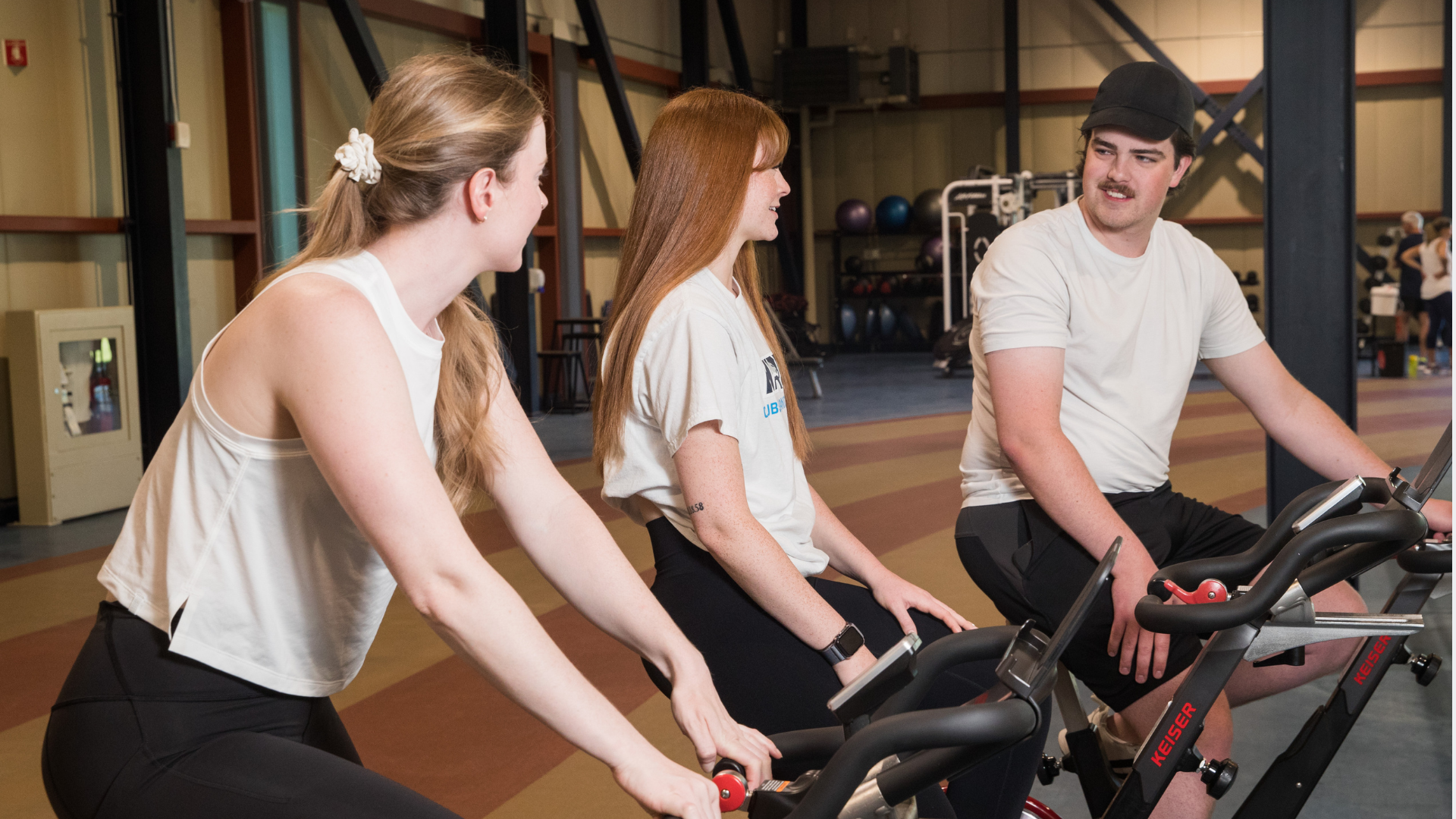 Three young adults sit on stationary bikes in a gym, smiling and chatting during a workout. Exercise equipment and fitness balls are visible in the background.