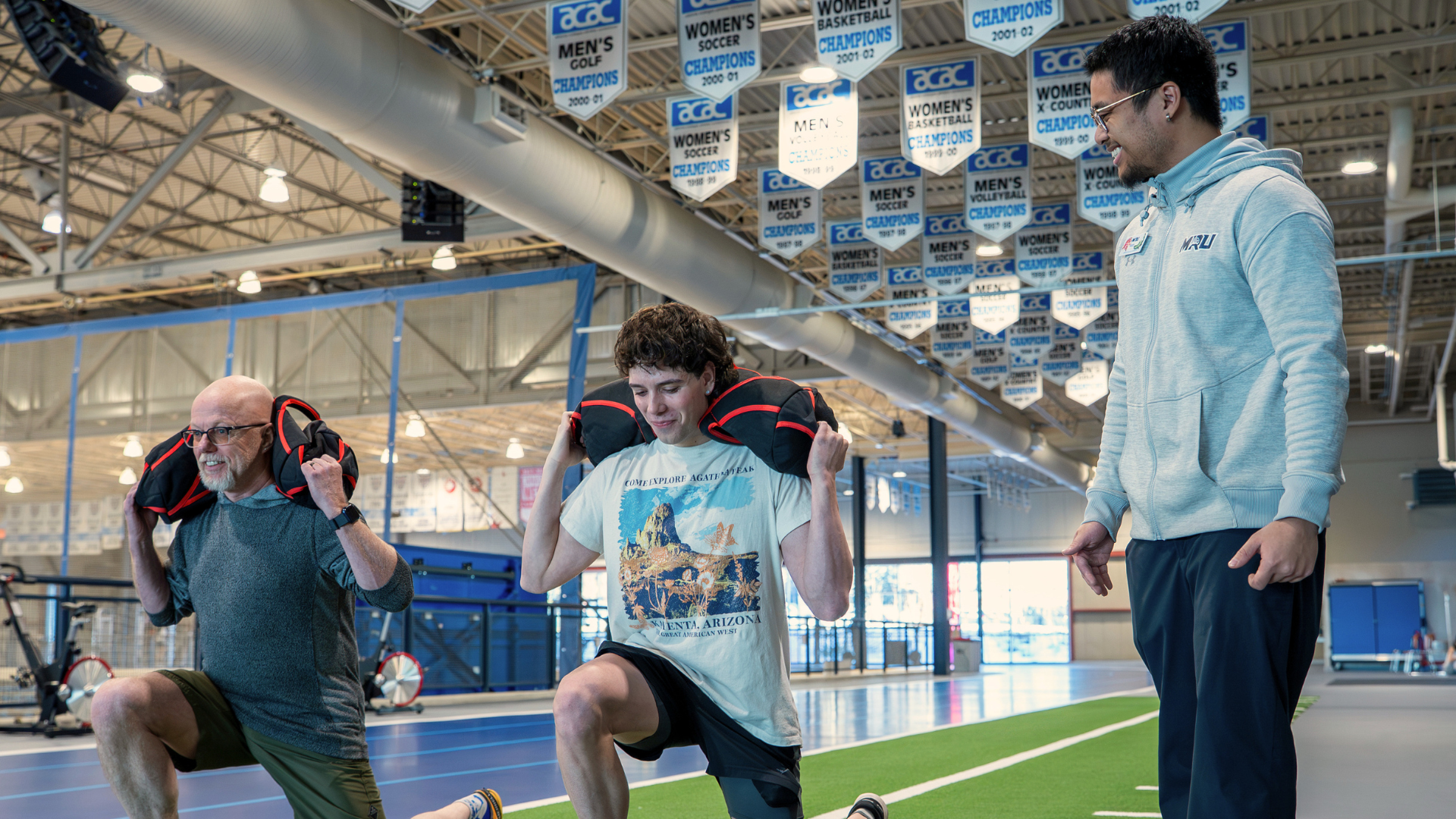  A fitness instructor supervises two participants performing weighted walking lunges on an indoor track inside a university recreation facility.