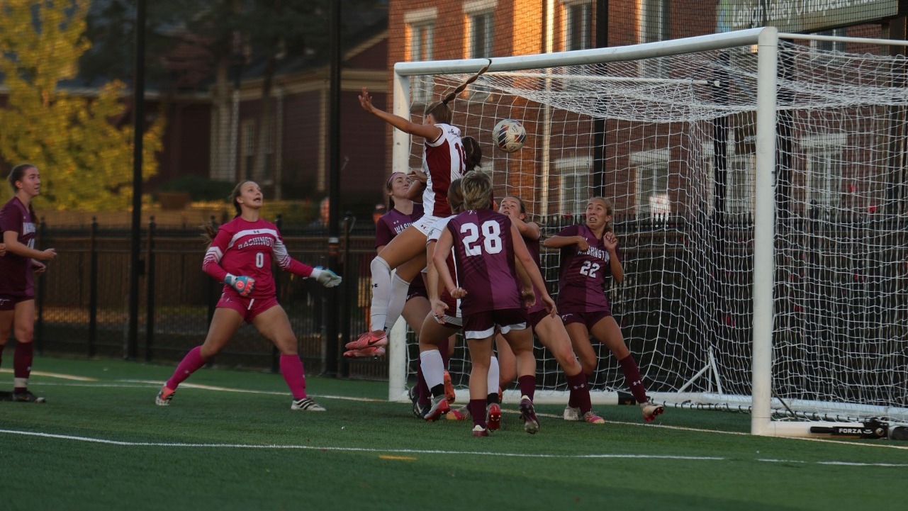 A crowd of soccer players in maroon jerseys surround one with a white jersey