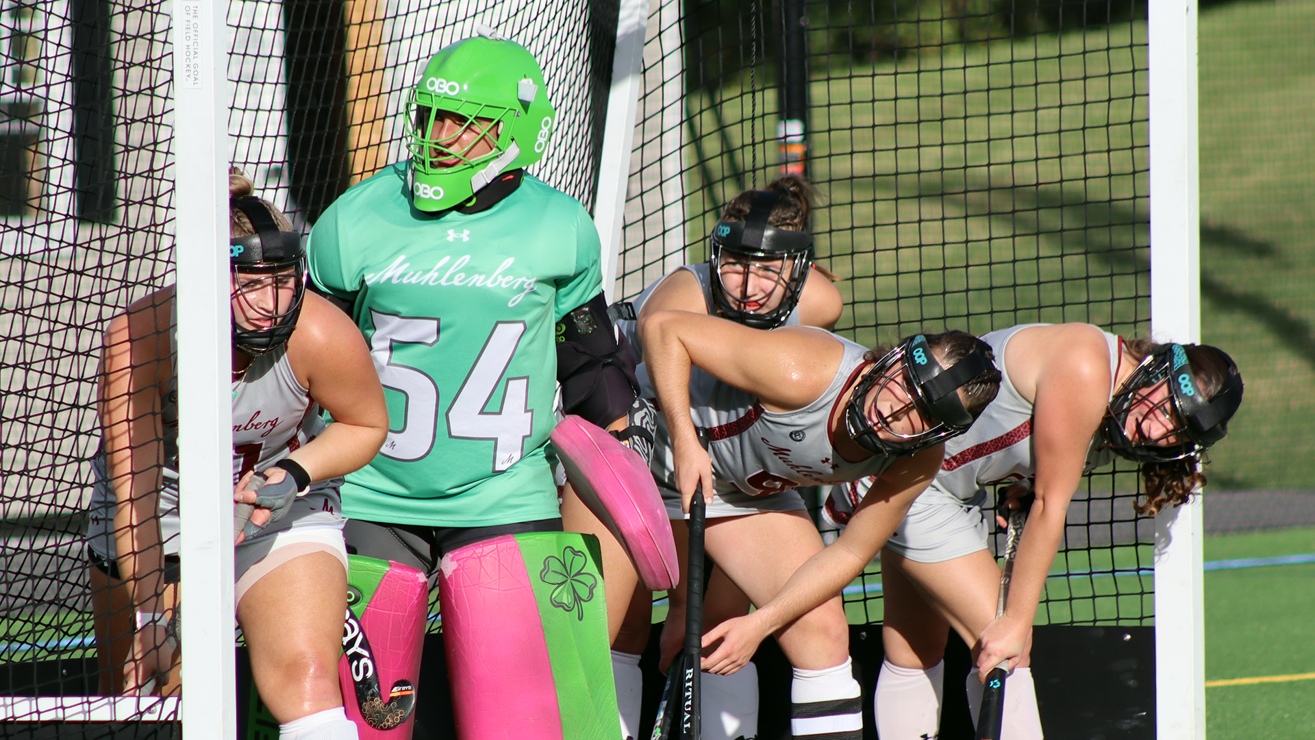 a goalie in a green jersey and field hockey players in grey jerseys stand in the cage