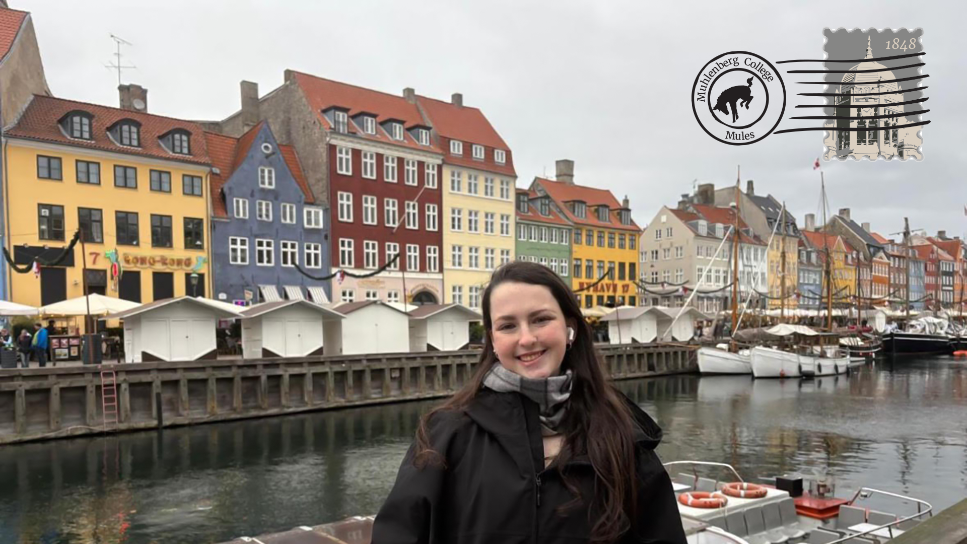 A young woman in a black coat stands in front of a waterway with boats and colorful buildings behind her